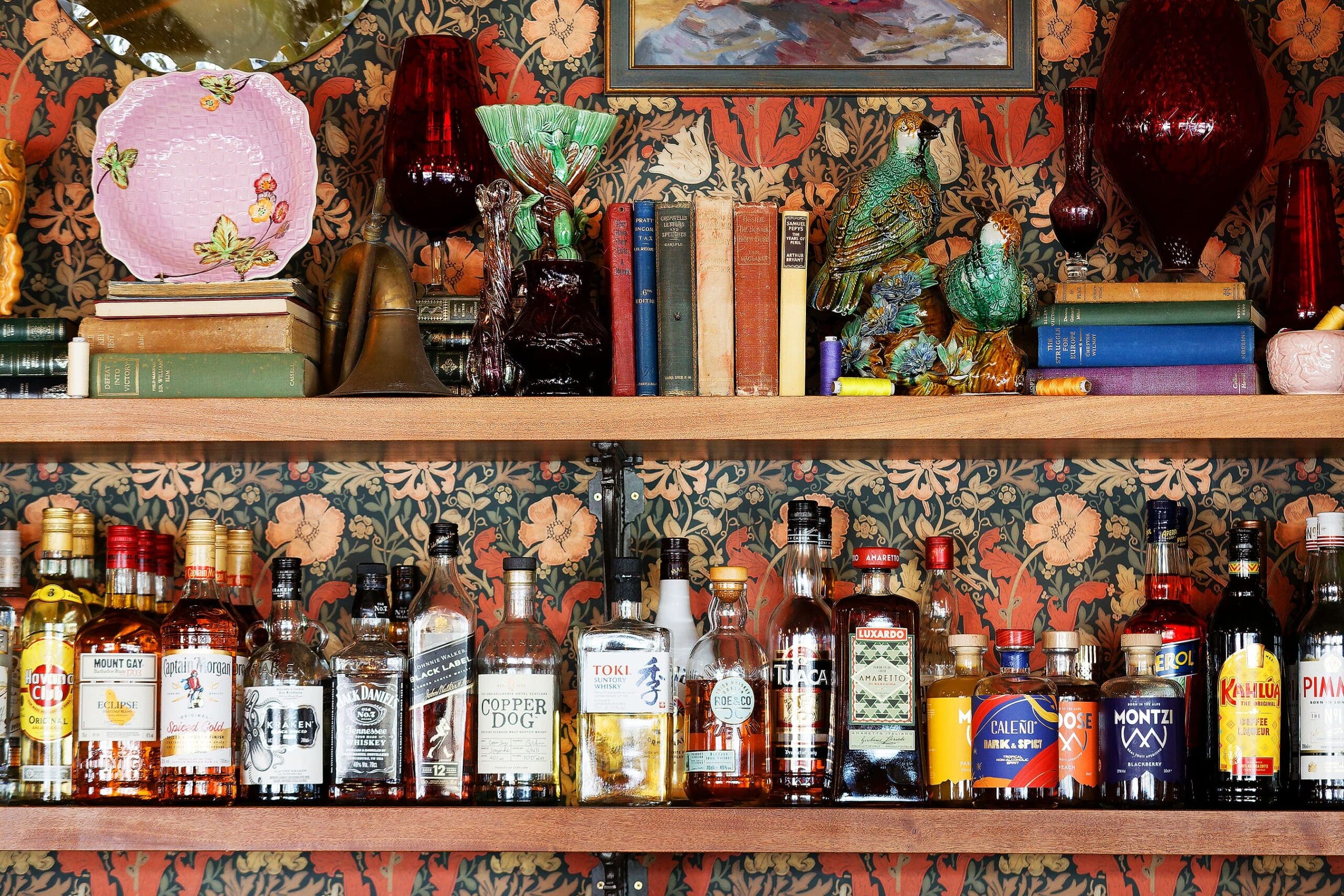 A wooden shelf against floral wallpaper holds various liquor bottles, glassware, books, a ceramic plate, and decorative figurines—including birds, a dog, and a small Juno statue. The bottom shelf displays assorted spirits and liqueurs.