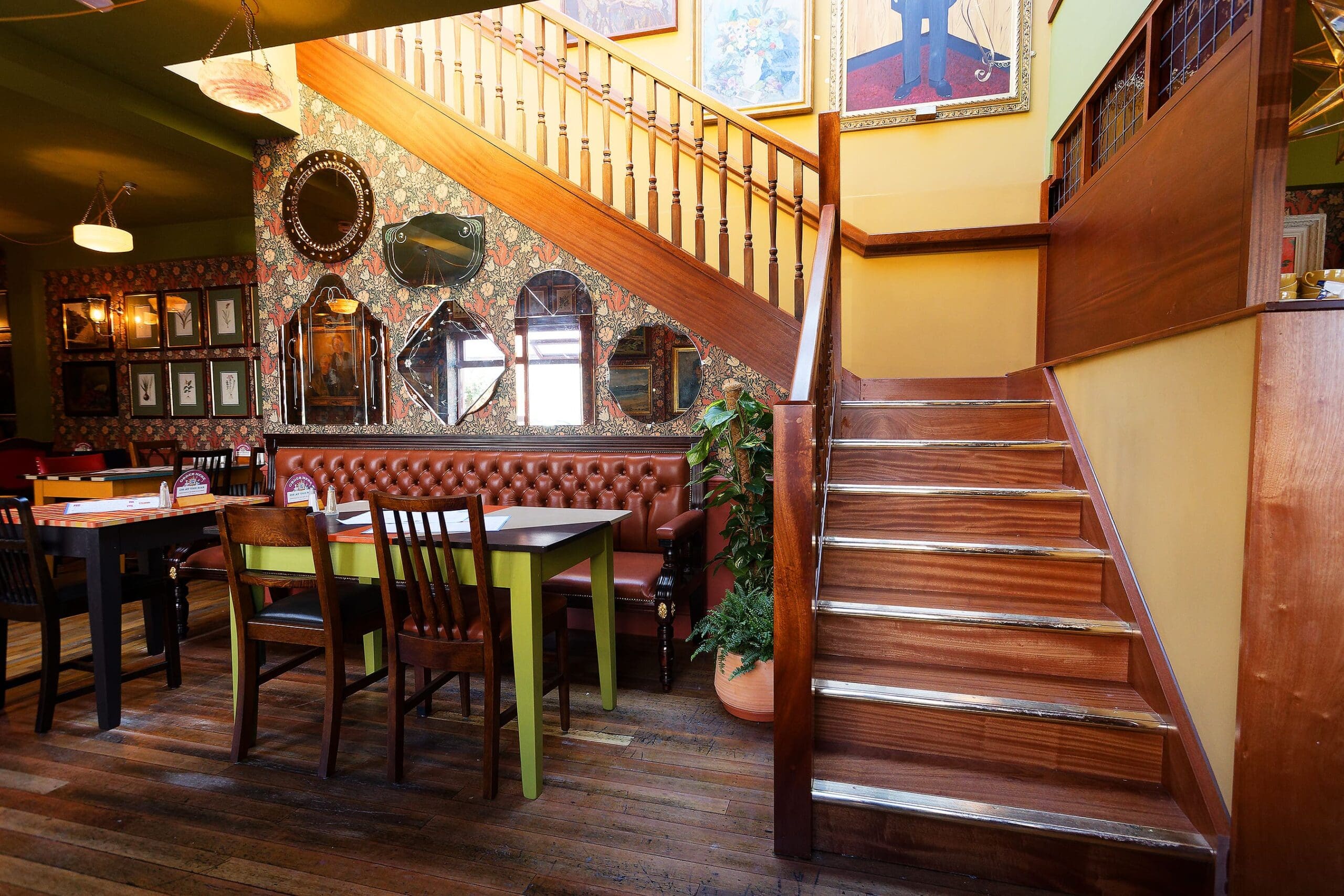 A cozy restaurant interior at Juno features wooden stairs, vintage wallpaper, framed mirrors and art, a brown tufted bench, and several tables and chairs on a wooden floor. Warm lighting creates an inviting atmosphere.