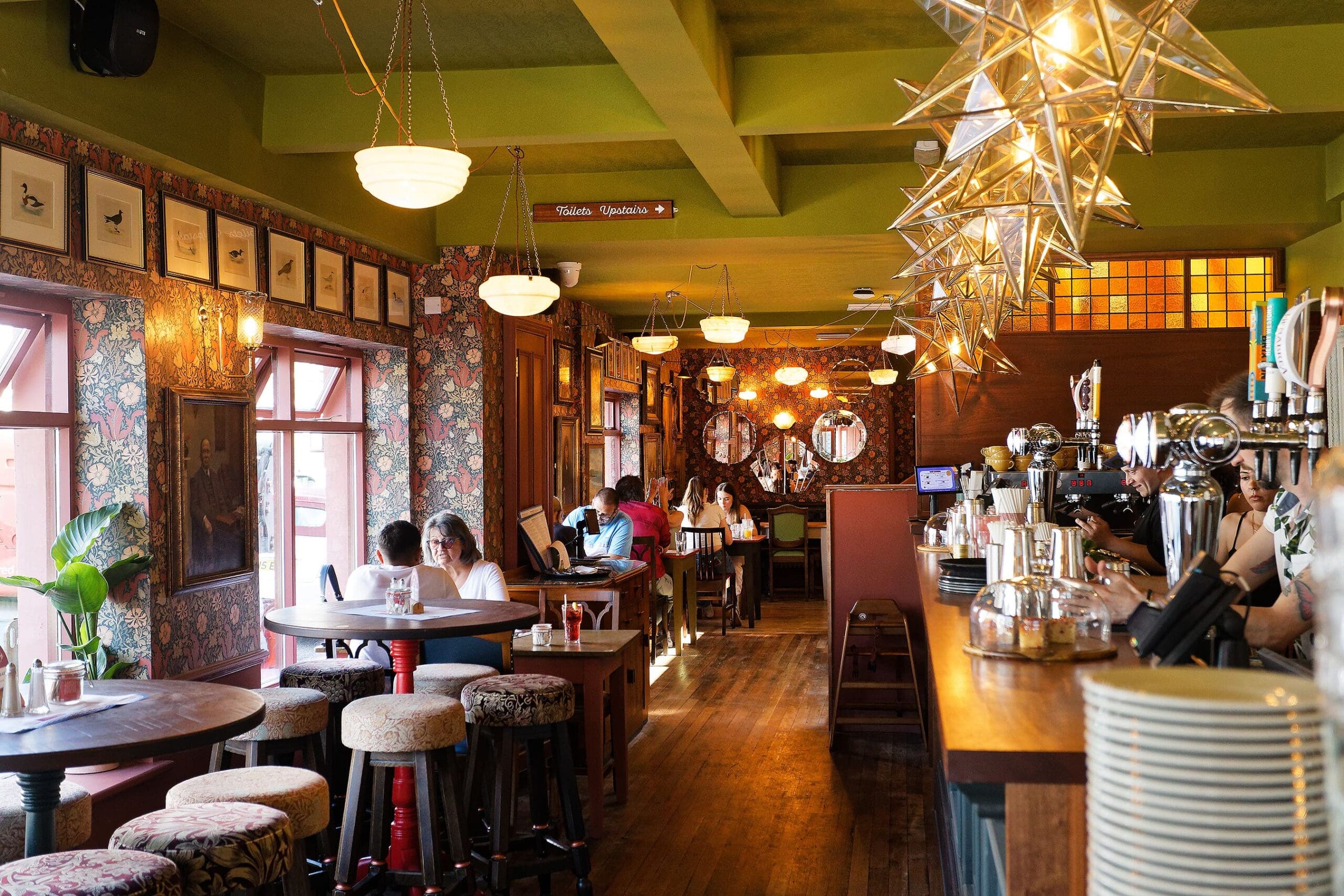 A cozy, vibrant bar named Juno, with patterned wallpaper, eclectic lighting, a long wooden counter, bar stools, and people sitting at tables by the windows, enjoying drinks and conversation.