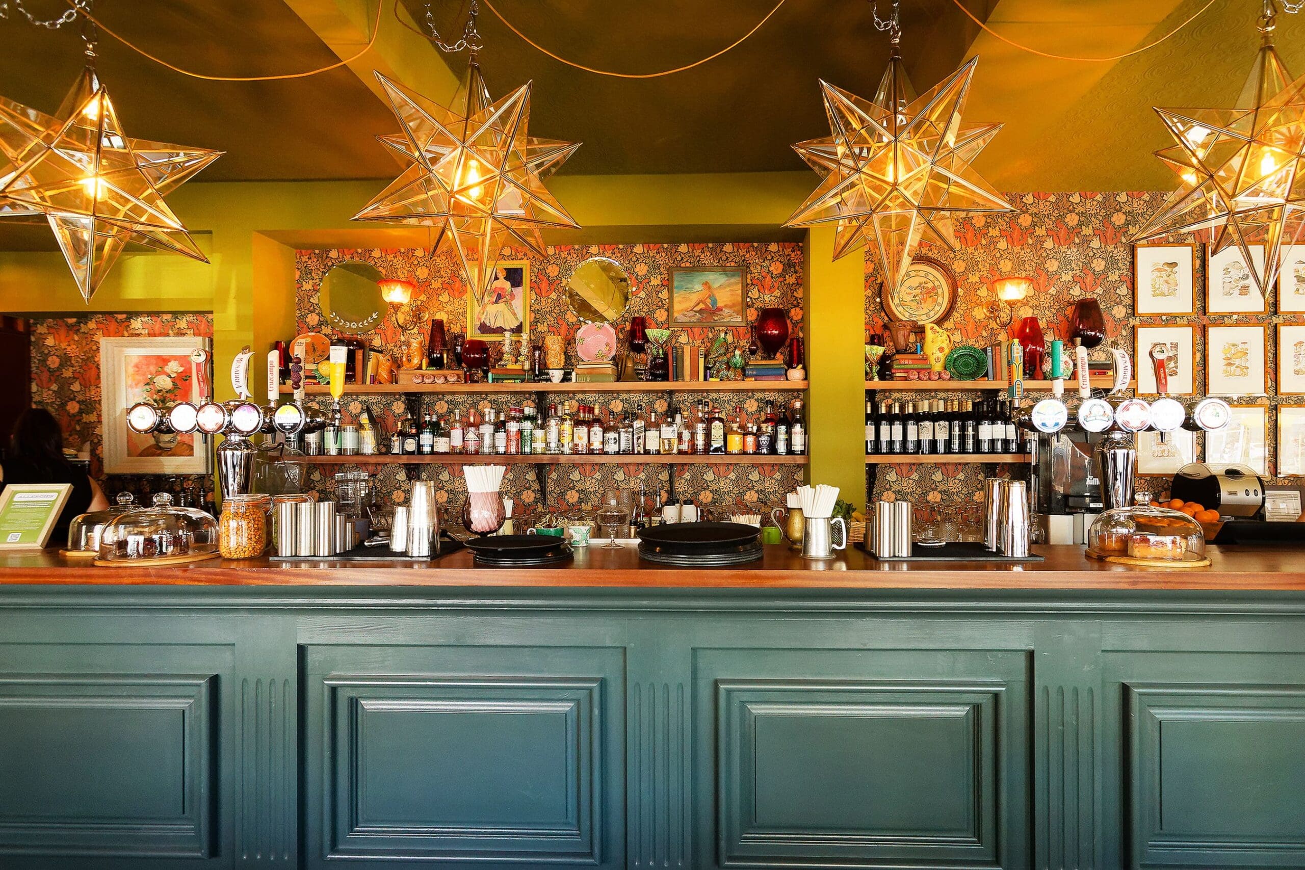 A colorful bar with a green counter, patterned wallpaper, shelves lined with bottles, and three large Juno star-shaped pendant lights hanging from the ceiling. The decor is eclectic and vibrant.