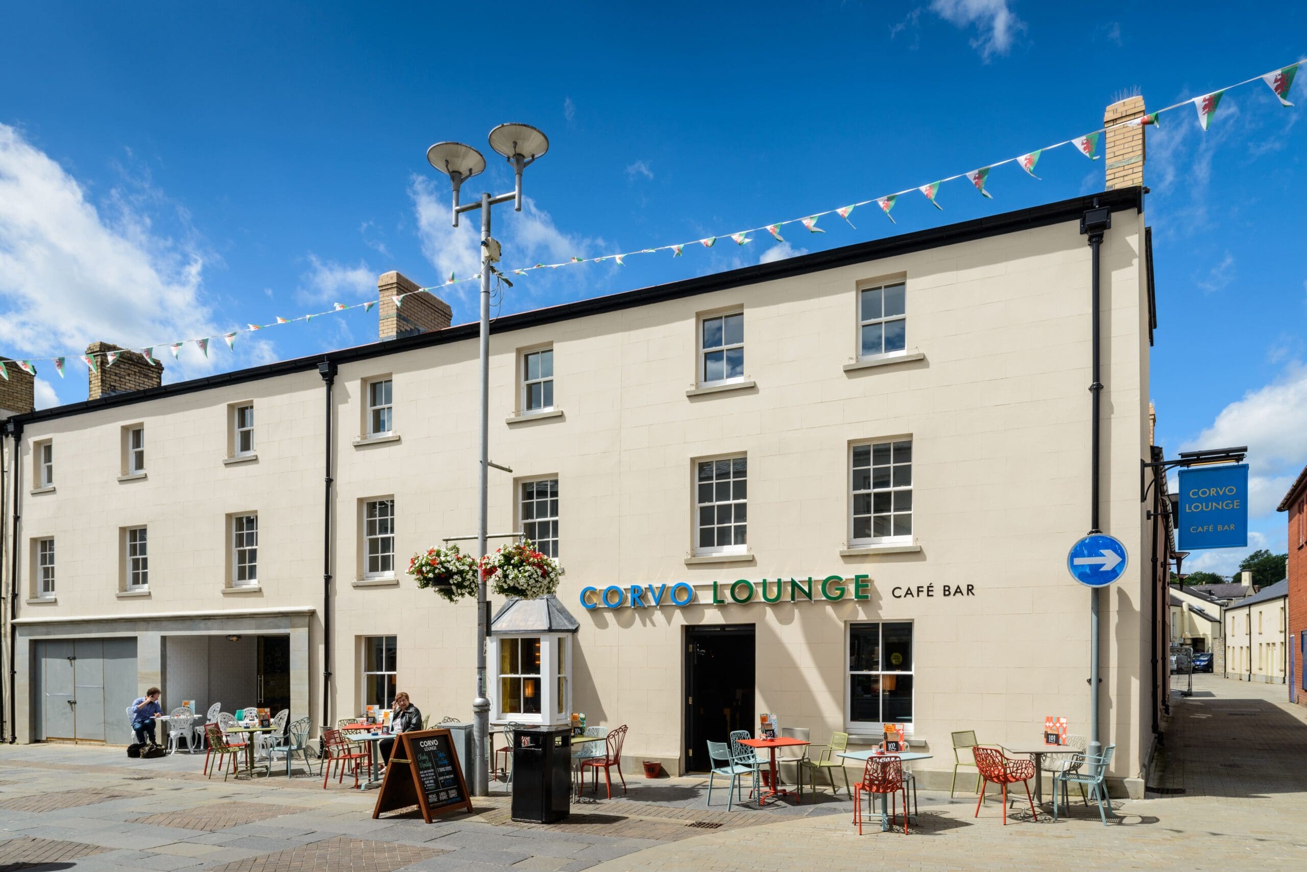 A bright, sunny day outside Corvo Lounge café bar; tables and chairs are set up on the pavement, with a few people seated. Colorful bunting hangs above the cream-colored Corvo building, creating a welcoming atmosphere.