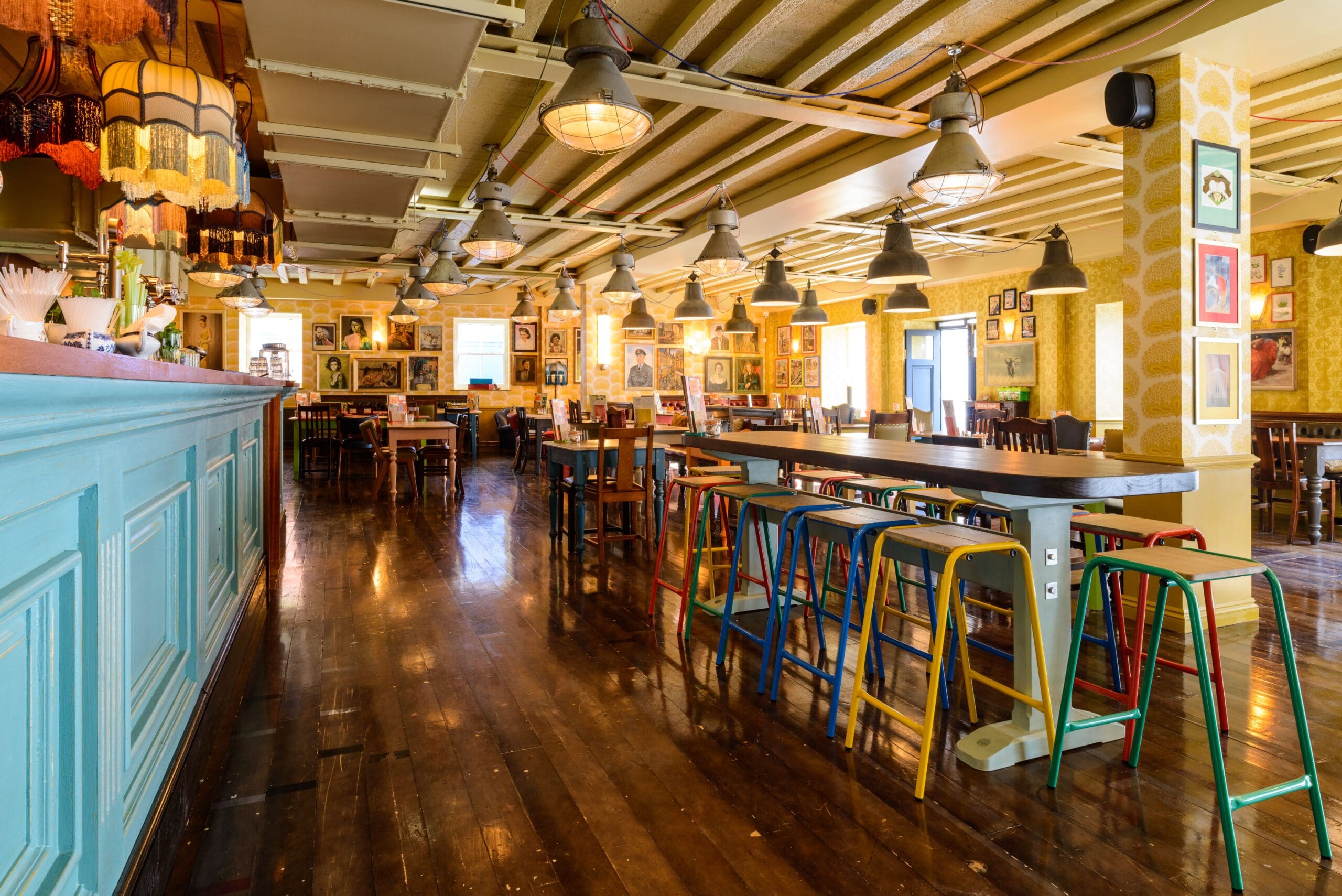 A brightly lit restaurant with colorful stools around high tables, wooden floors, pendant lighting, and walls adorned with art. The turquoise Corvo counter is on the left.