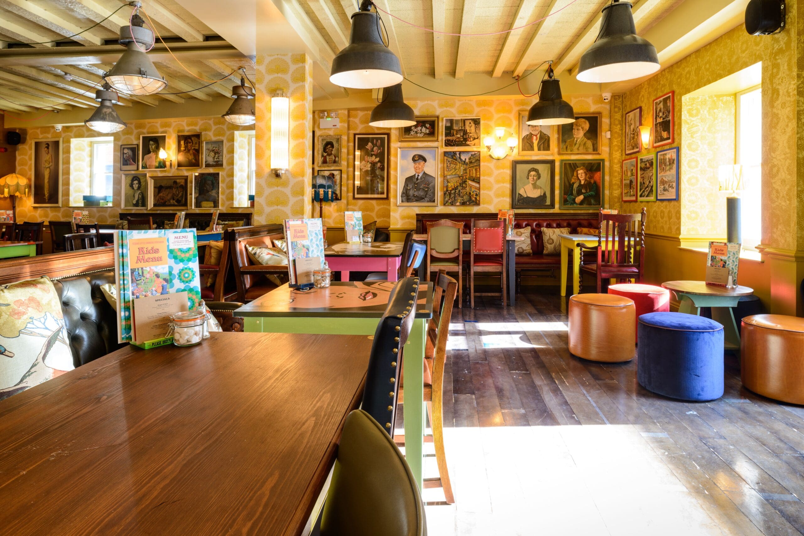 A cozy, brightly lit cafe with wooden tables, colorful chairs and stools, wall art with various portraits including Corvo, yellow wallpaper, and large windows letting in sunlight. Hanging pendant lights create a warm and inviting atmosphere.