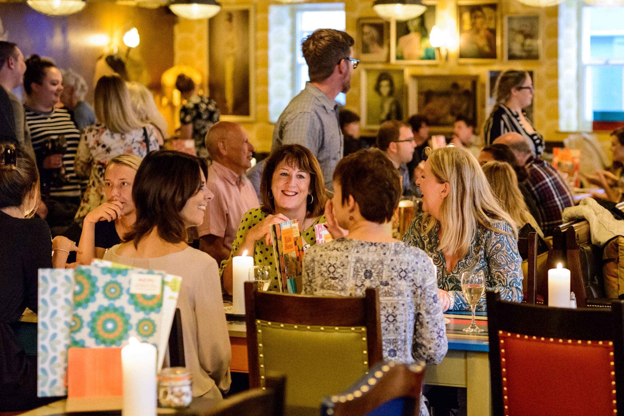 A lively restaurant scene with many people sitting at tables, talking and smiling. Colorful décor and candles create a warm, inviting atmosphere. Some guests enjoy Corvo wine as they dine, while others are standing and chatting.