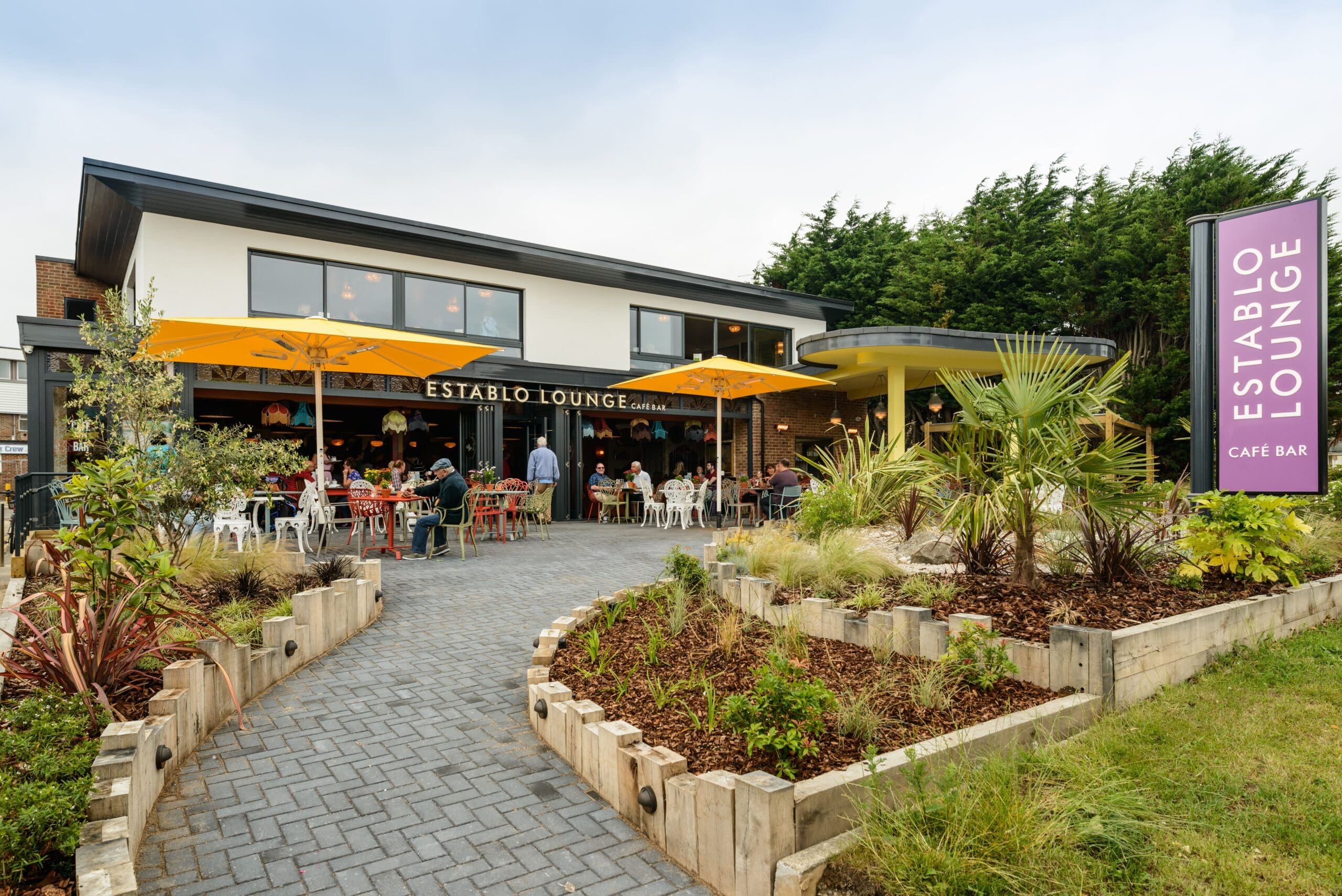 Outdoor view of Establo Lounge café bar with a garden pathway, colorful umbrellas, tables with people dining, and a purple Establo sign. Landscaped plants frame the modern two-story building in the background.