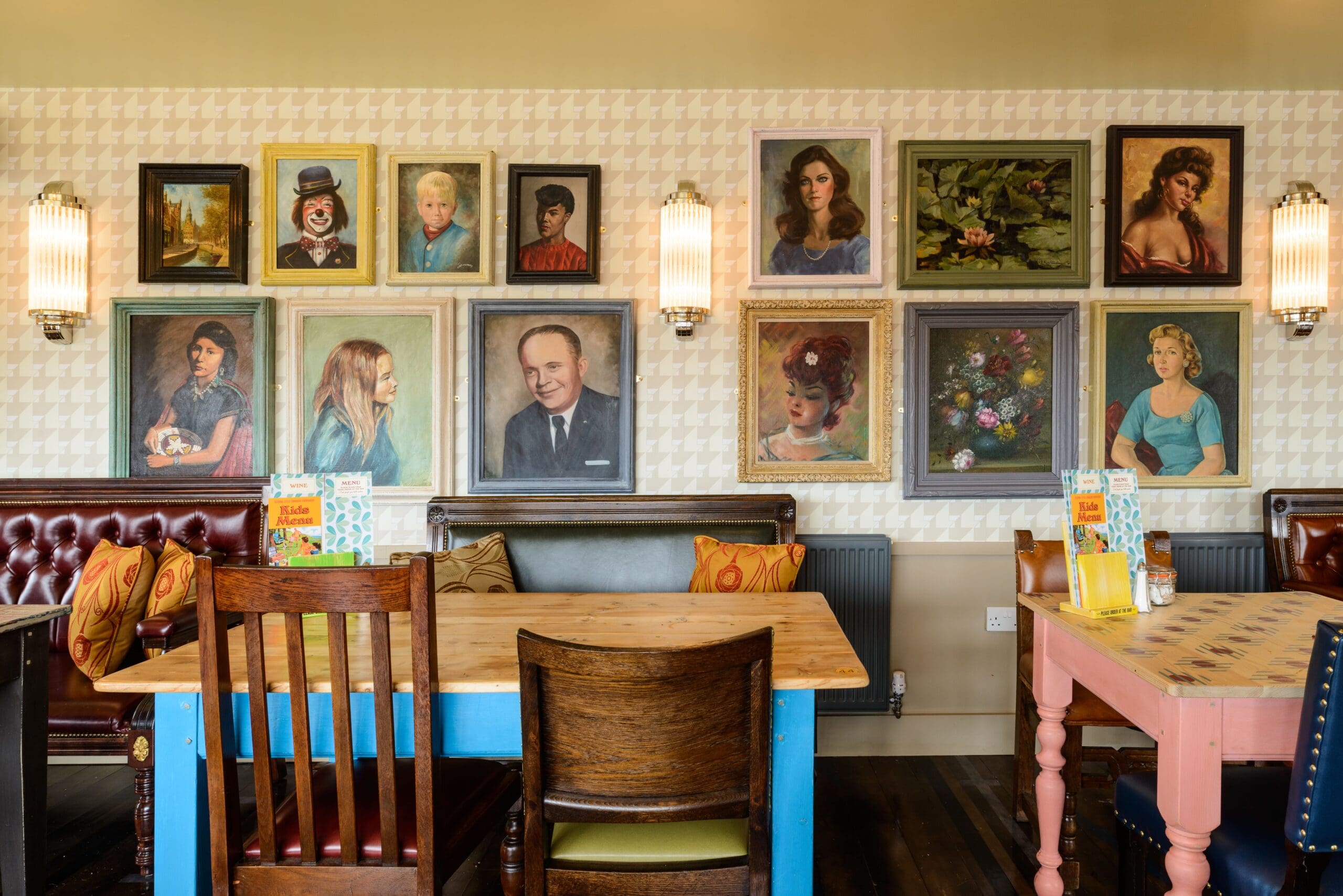 A cozy cafe, Establo, with wooden tables and colorful chairs features a wall decorated with framed vintage portraits and still life paintings. Two wall sconces provide warm lighting, while menus and condiments sit ready on the tables.