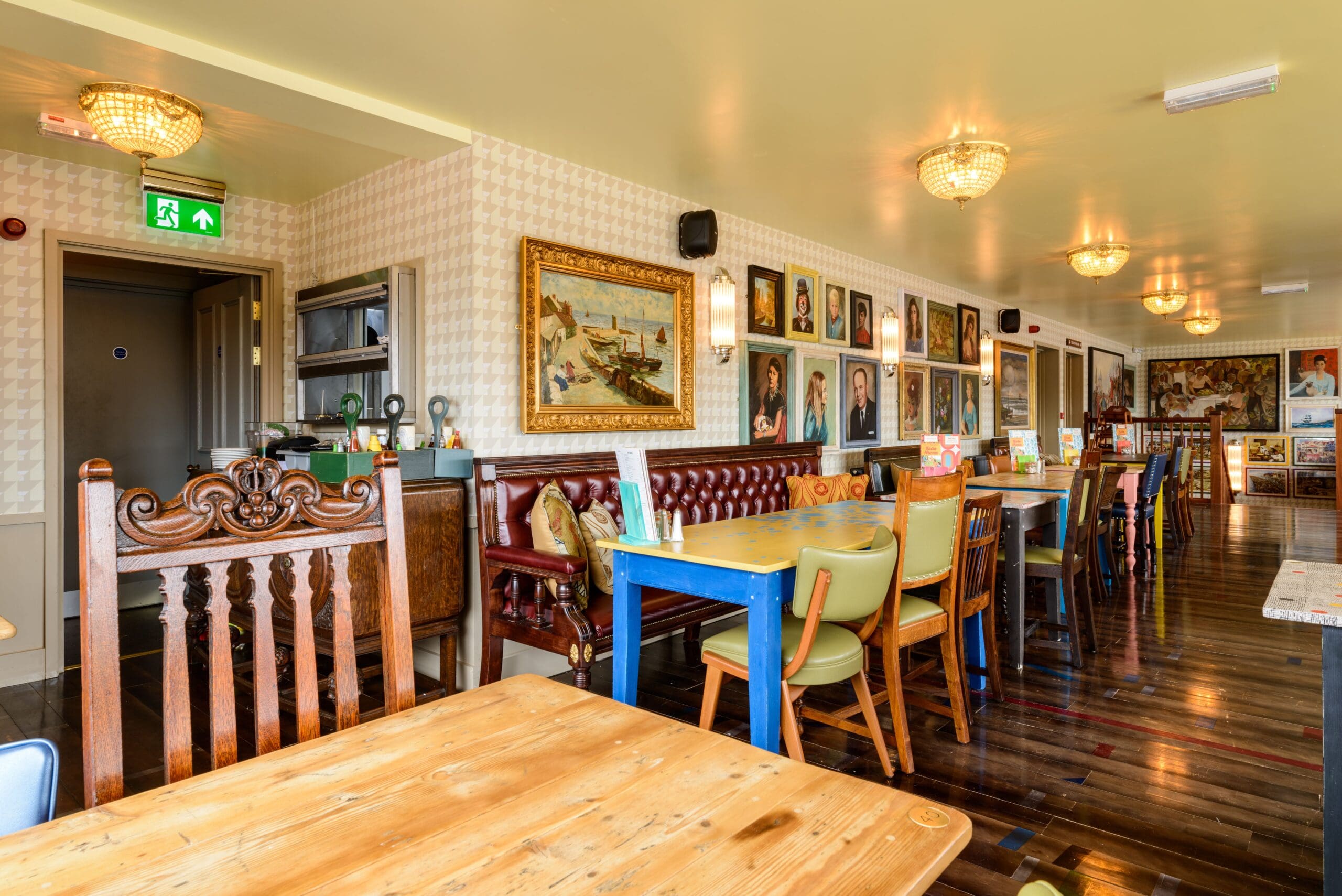 A bright, eclectic Establo restaurant interior with wooden tables, mismatched chairs, a brown leather bench, chandelier lights, and walls adorned with various framed paintings and portraits.
