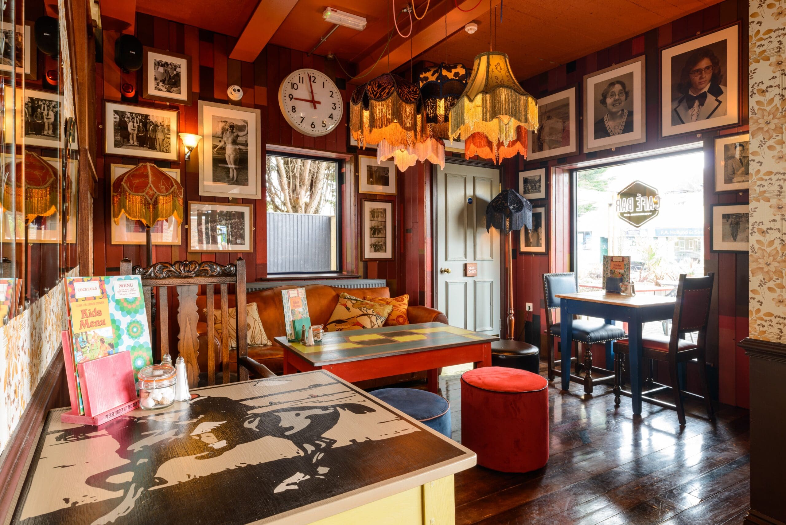 Cozy Establo vintage-style café interior with eclectic lampshades, framed photos on red paneled walls, mismatched tables and seating, and large windows letting in natural light. A clock is mounted above the door.