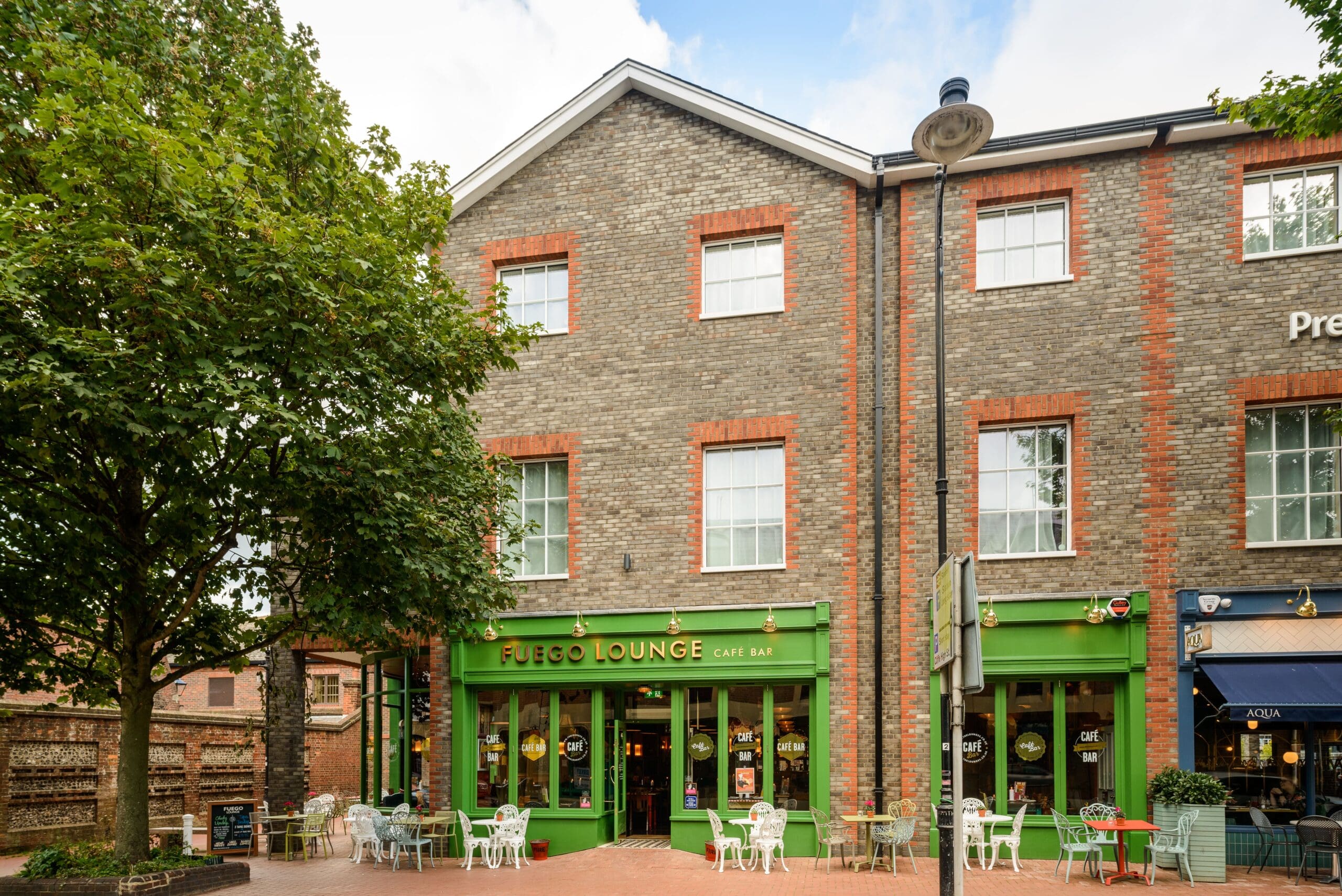 Street view of a modern brick building with the green-trimmed café bar Fuego Lounge; outdoor tables and chairs are set up on the sidewalk, and a tree stands to the left, inviting you to experience Fuego’s lively atmosphere.