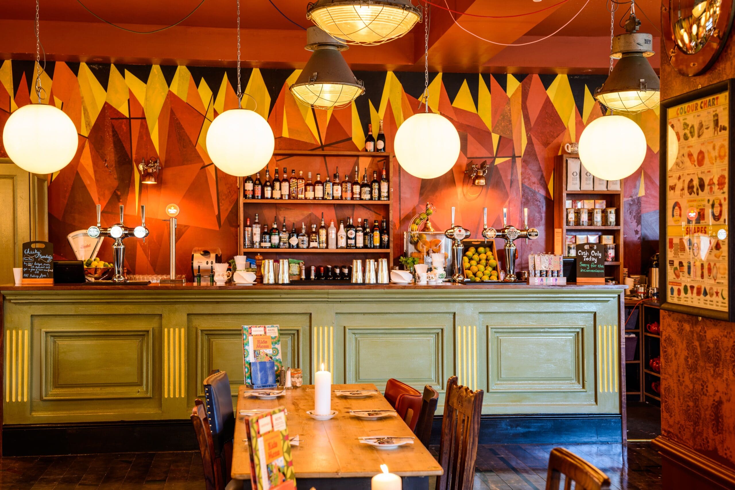 A cozy bar with a retro vibe, Fuego features a green wooden counter, three round hanging lights, colorful geometric wall art, bottles on shelves, and a wooden table with menus and chairs in the foreground.