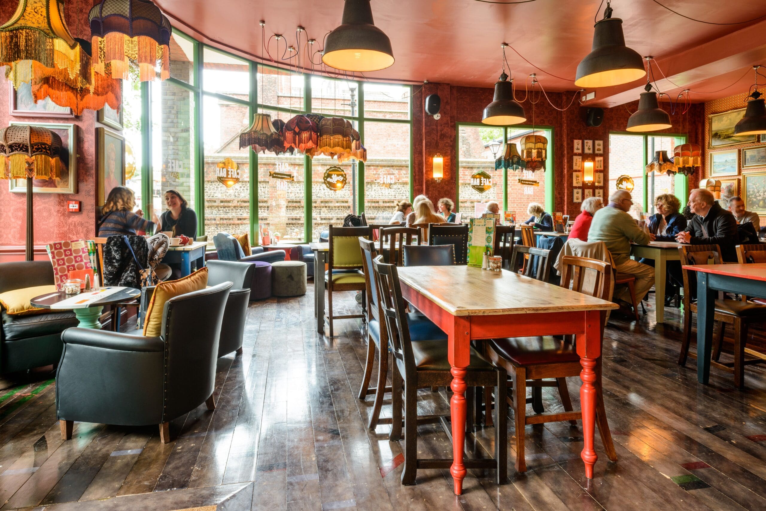 A cozy, eclectic café with colorful lampshades, mismatched furniture, and large windows. Groups of people are sitting and chatting at various tables, adding Fuego to the warm, lively atmosphere.