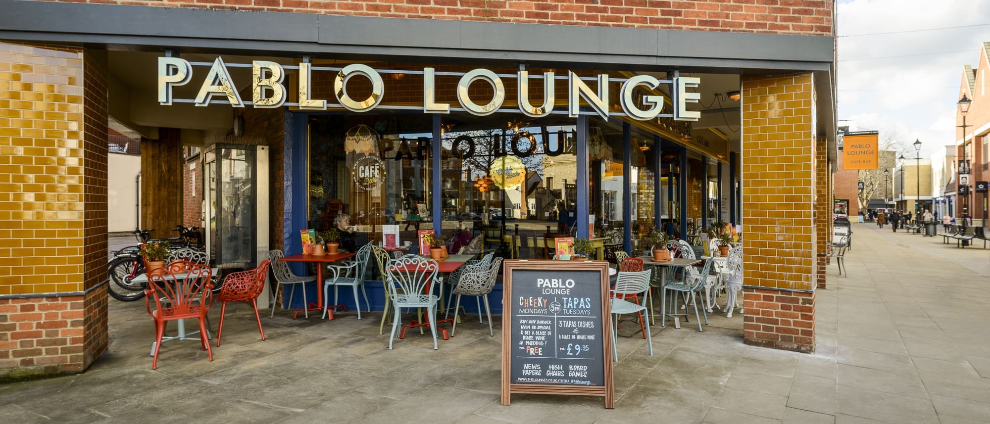 Outdoor seating at Pablo Lounge café with colorful chairs and tables, a large Pablo sign above the entrance, and a chalkboard menu on the sidewalk. The café offers a vibrant, welcoming atmosphere on a lively pedestrian street.