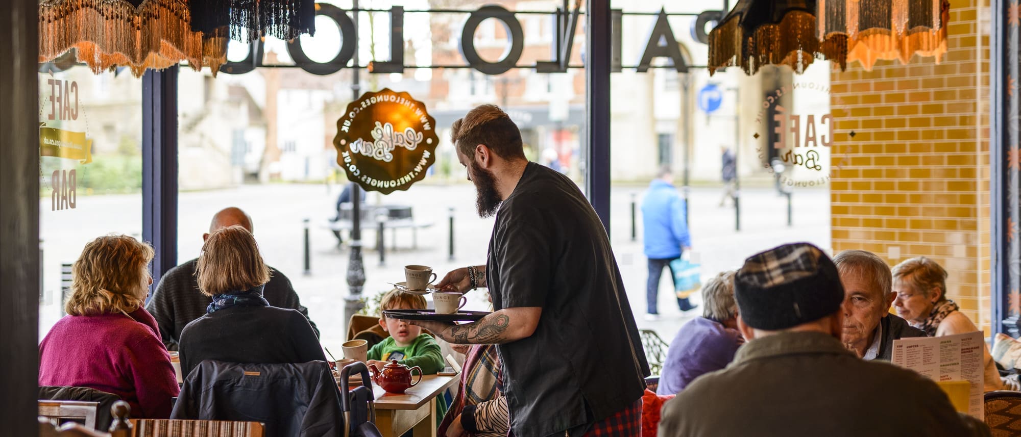 A busy café scene with people sitting at tables by large windows, chatting and drinking. Pablo, a bearded server, carries a tray with drinks, while others read menus or talk. Outside, a street and buildings are visible through the glass.