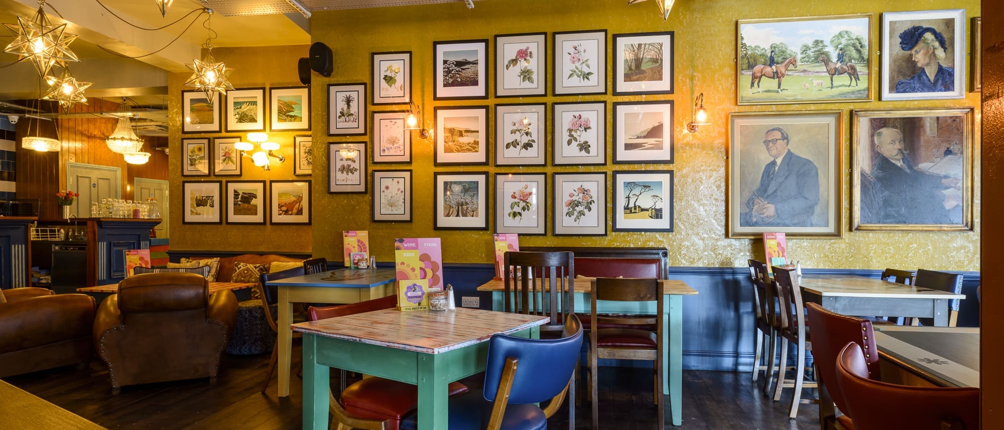 A cozy café with mismatched colorful chairs and tables, decorated with numerous framed pictures and portraits—some inspired by Pablo—on the walls, warm lighting, and menus placed on each table.