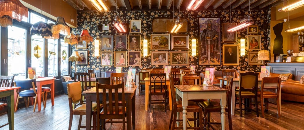 A cozy, eclectic cafe with mismatched chairs and tables, floral wallpaper, and walls covered in framed portraits and Benito-inspired art. Warm pendant lights and natural sunlight create a welcoming atmosphere.