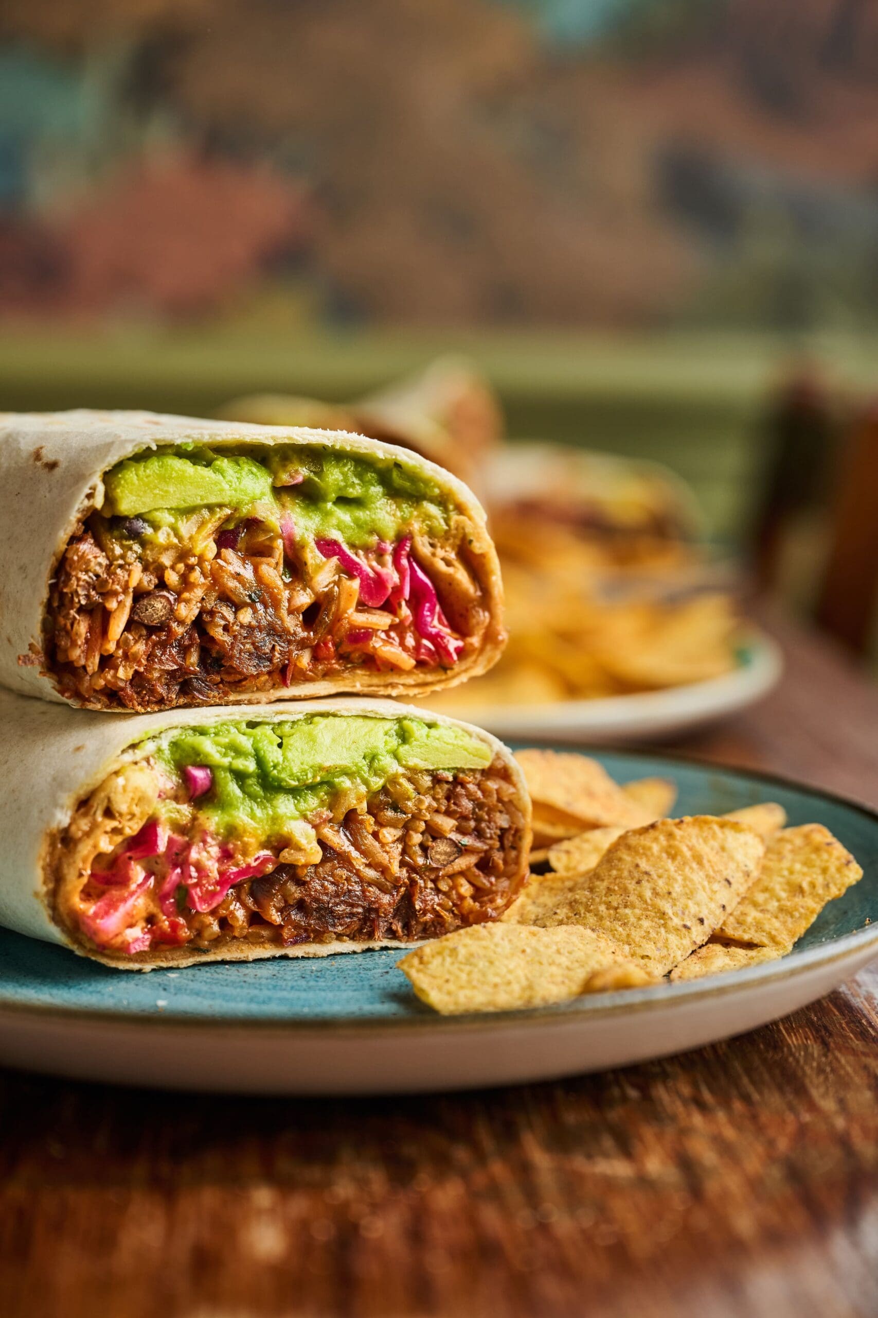 A burrito cut in half, showing shredded meat, guacamole, and pickled onions inside, is served Torino-style on a plate with tortilla chips.