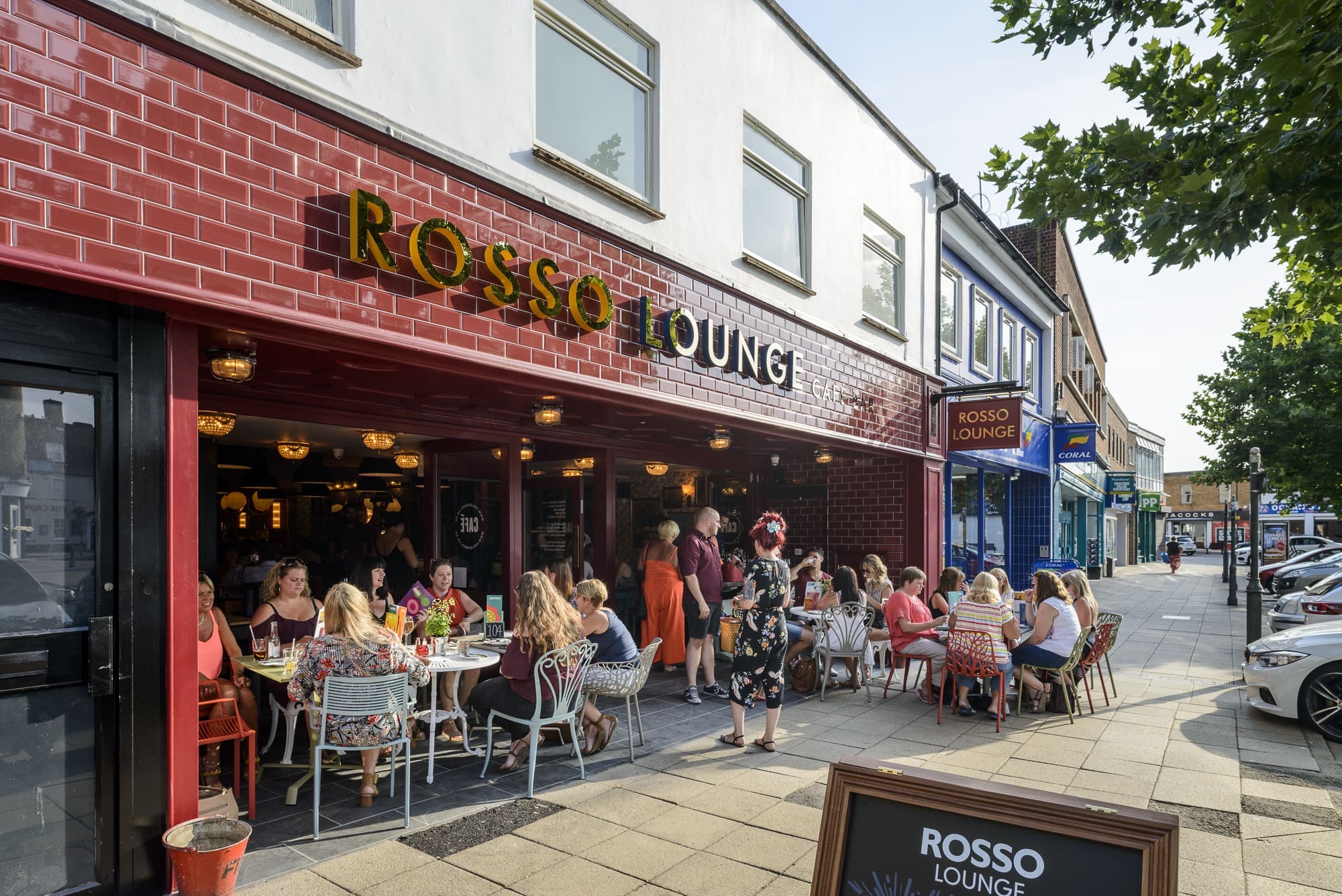 People sit at outdoor tables eating and chatting outside Rosso, a restaurant with a red tiled facade. Other shops and parked cars line the street, while trees provide shade on the sidewalk.