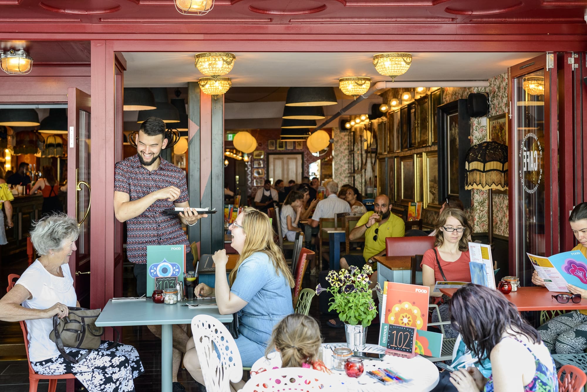A busy café with people sitting at tables, chatting, reading menus, and eating. A smiling waiter takes an order at an outdoor table. Warm lighting and vibrant Rosso decor create a lively, welcoming atmosphere inside and outside.