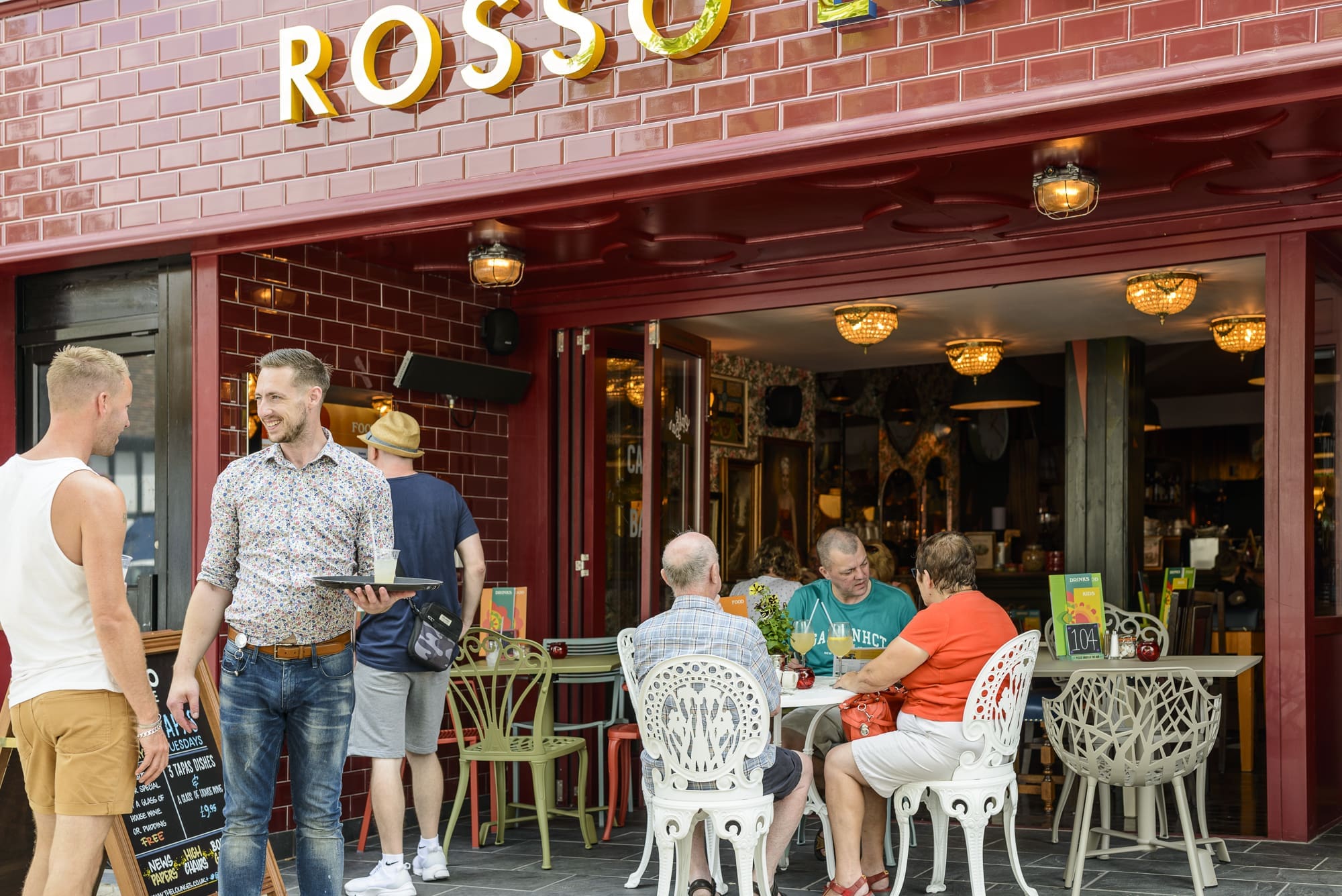 Outdoor seating at Rosso features red tiles, with people at white tables enjoying food and drinks. A waiter chats with a standing guest while others in casual summer attire relax under the "ROSSO" sign above.