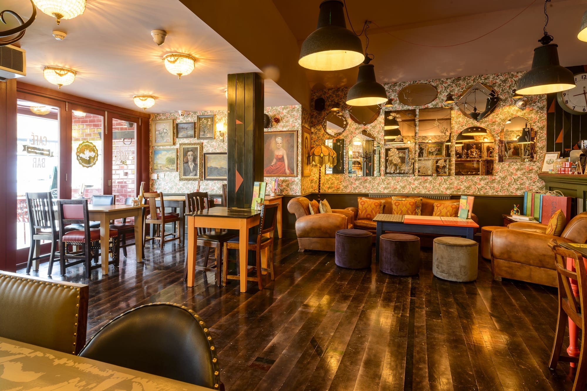 A cozy, warmly lit café with wooden floors, eclectic furniture, Rosso accents, wall art, decorative mirrors, books, and vintage-style lighting creates a comfortable and inviting atmosphere.