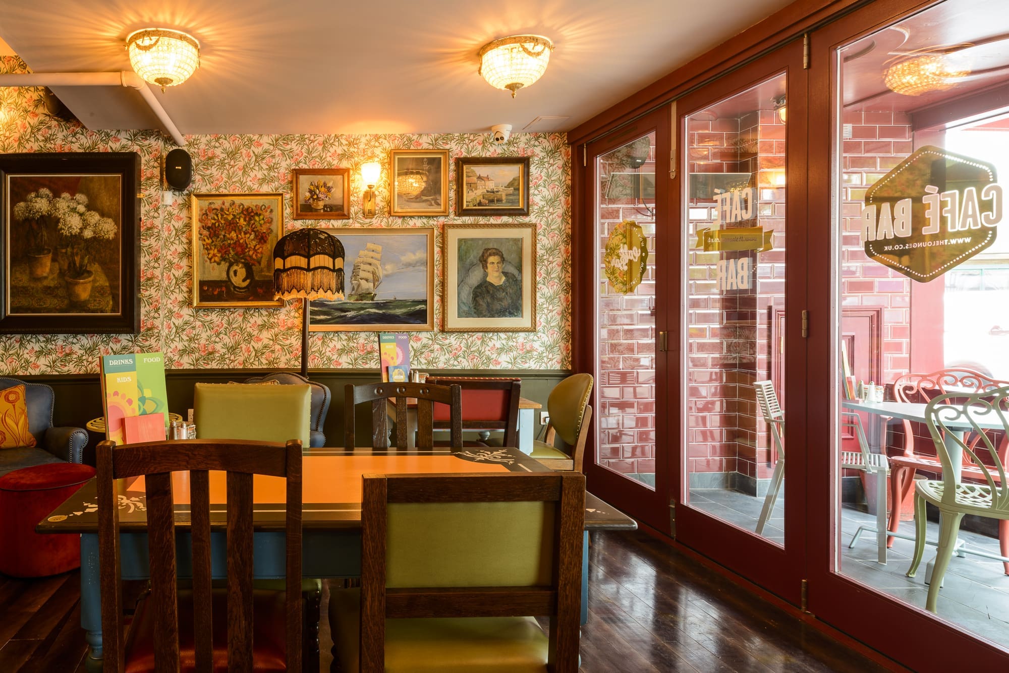 A cozy café interior with colorful chairs, a wooden table, floral wallpaper, framed vintage paintings on the wall, warm lighting, and large Rosso-framed glass doors leading to an inviting outdoor seating area.