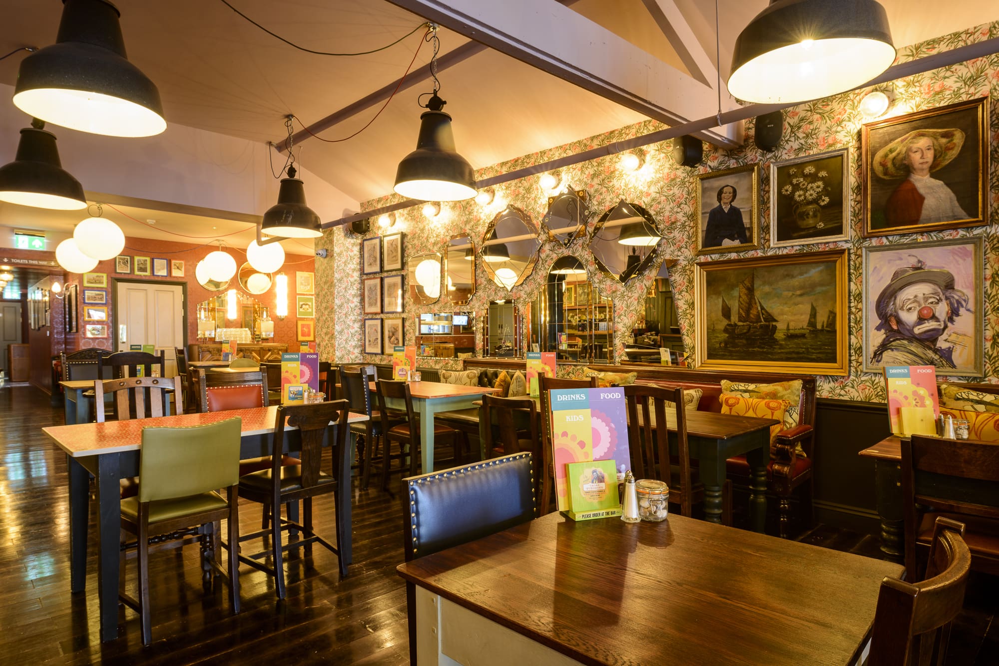 A cozy, eclectic restaurant interior with colorful chairs, wooden tables, and Rosso accents. Hanging lights illuminate walls adorned with mirrors, framed paintings, and portraits. Menus sit on tables in this warm, inviting atmosphere.