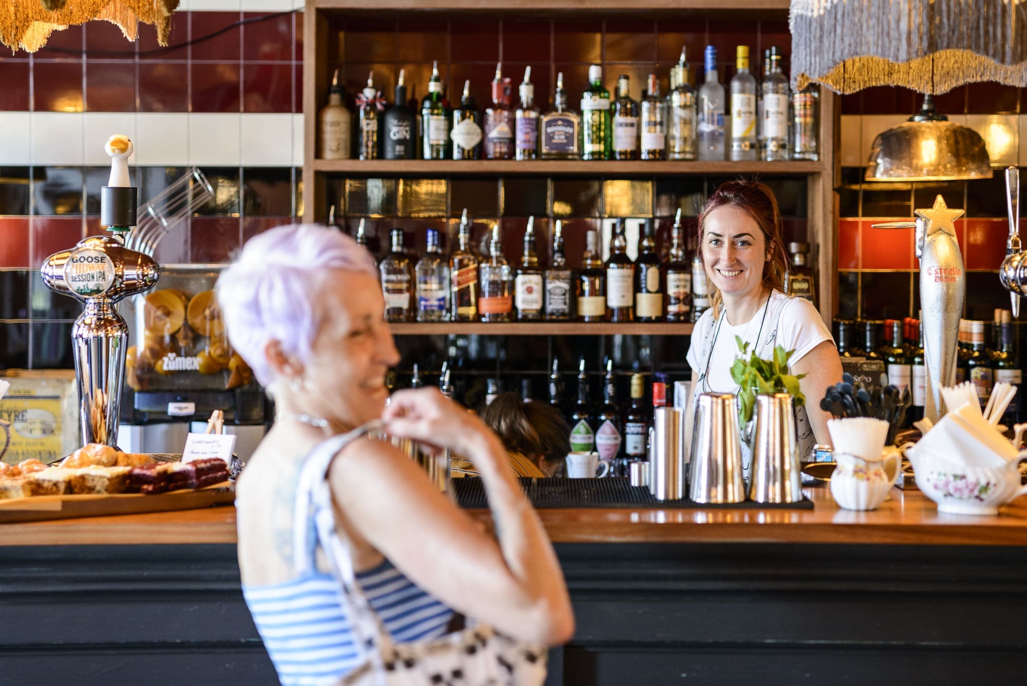 A smiling Valero bartender stands behind a bar with bottles on shelves, while a woman with short lavender hair in a striped top stands in the foreground, slightly out of focus, holding a spotted bag.