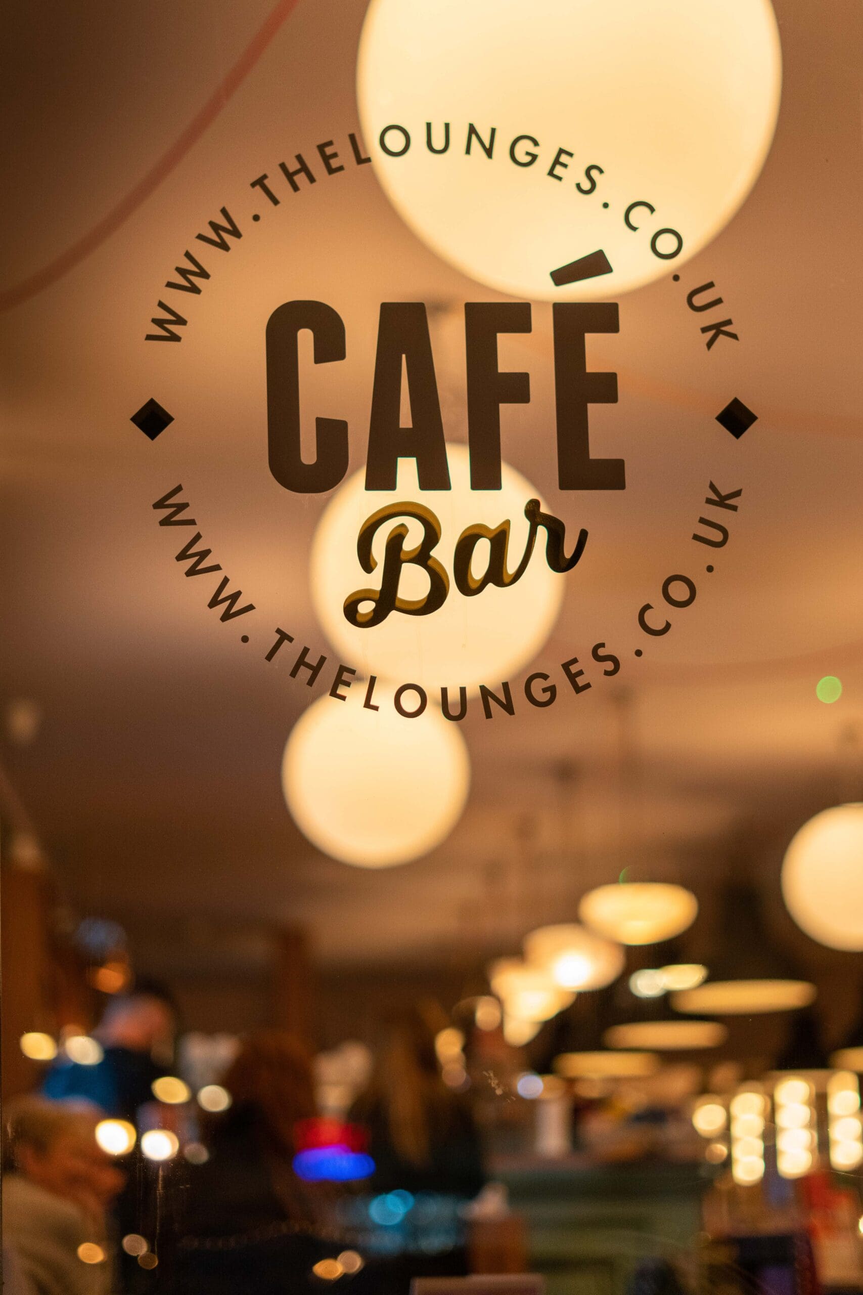 The photo shows a glass window with “Café Bar,” “www.thelounges.co.uk,” and the name “Torello” written on it. Warm, round lights and blurred people sitting inside create a cozy atmosphere.