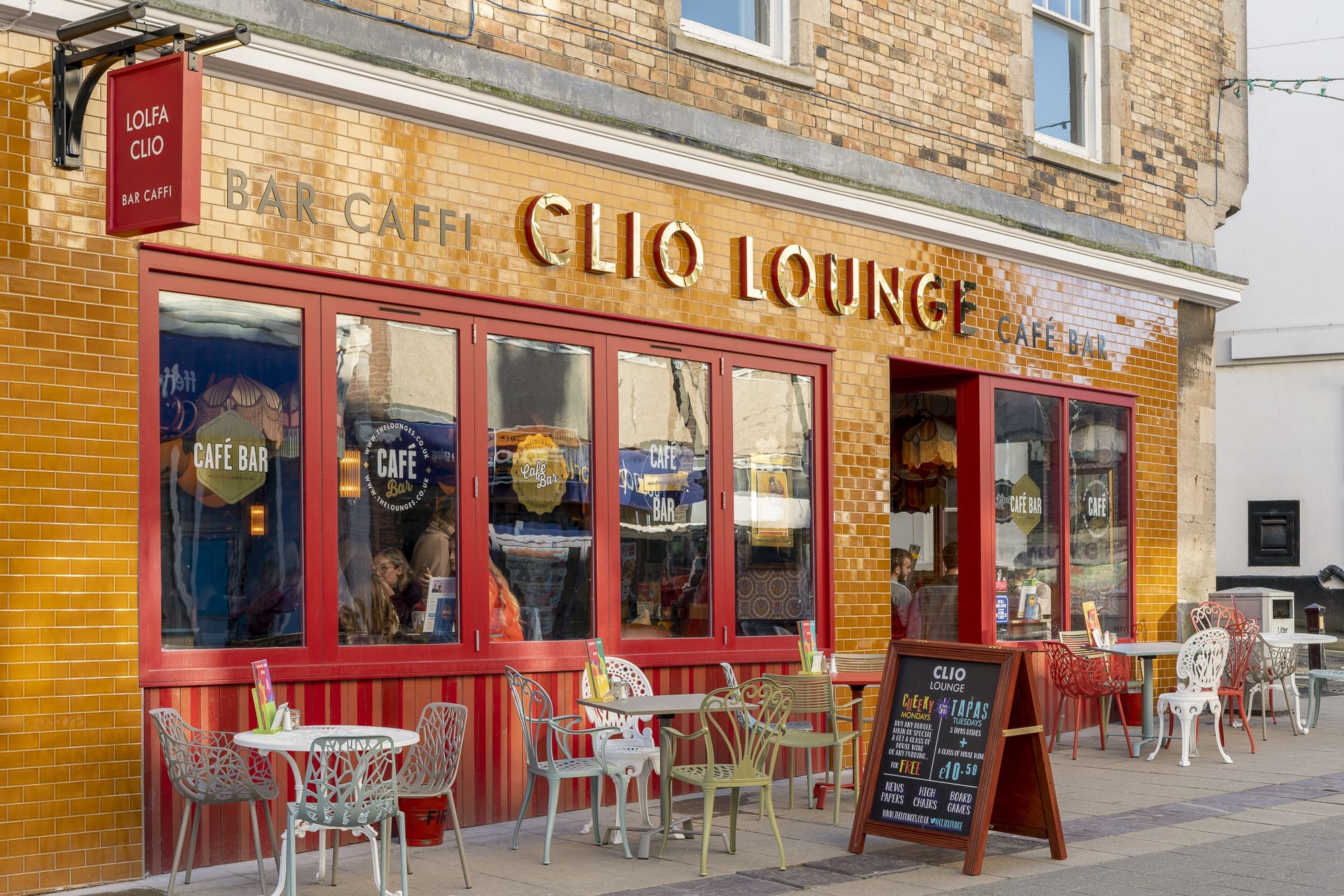 Street view of Clio Lounge café with red-framed windows and outdoor seating. Several tables and chairs are arranged on the pavement, a chalkboard menu stands near the entrance, and "Clio" signage tops the yellow-tiled building.