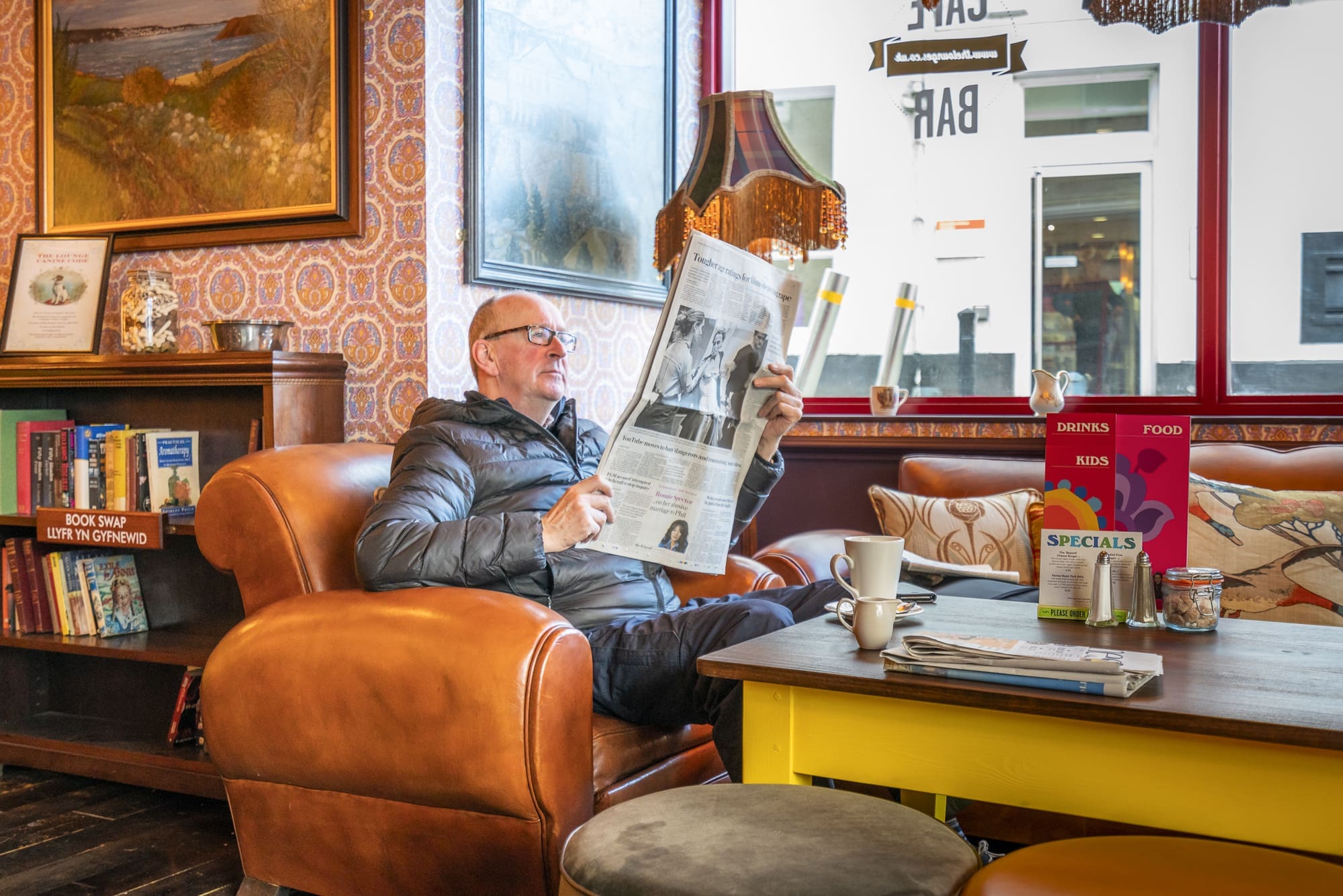 A man wearing glasses and a jacket sits in a cozy Clio café on a leather armchair, reading a newspaper. Books, a lamp, and menu signs are on nearby tables, with warm light and vintage decor creating a relaxed atmosphere.