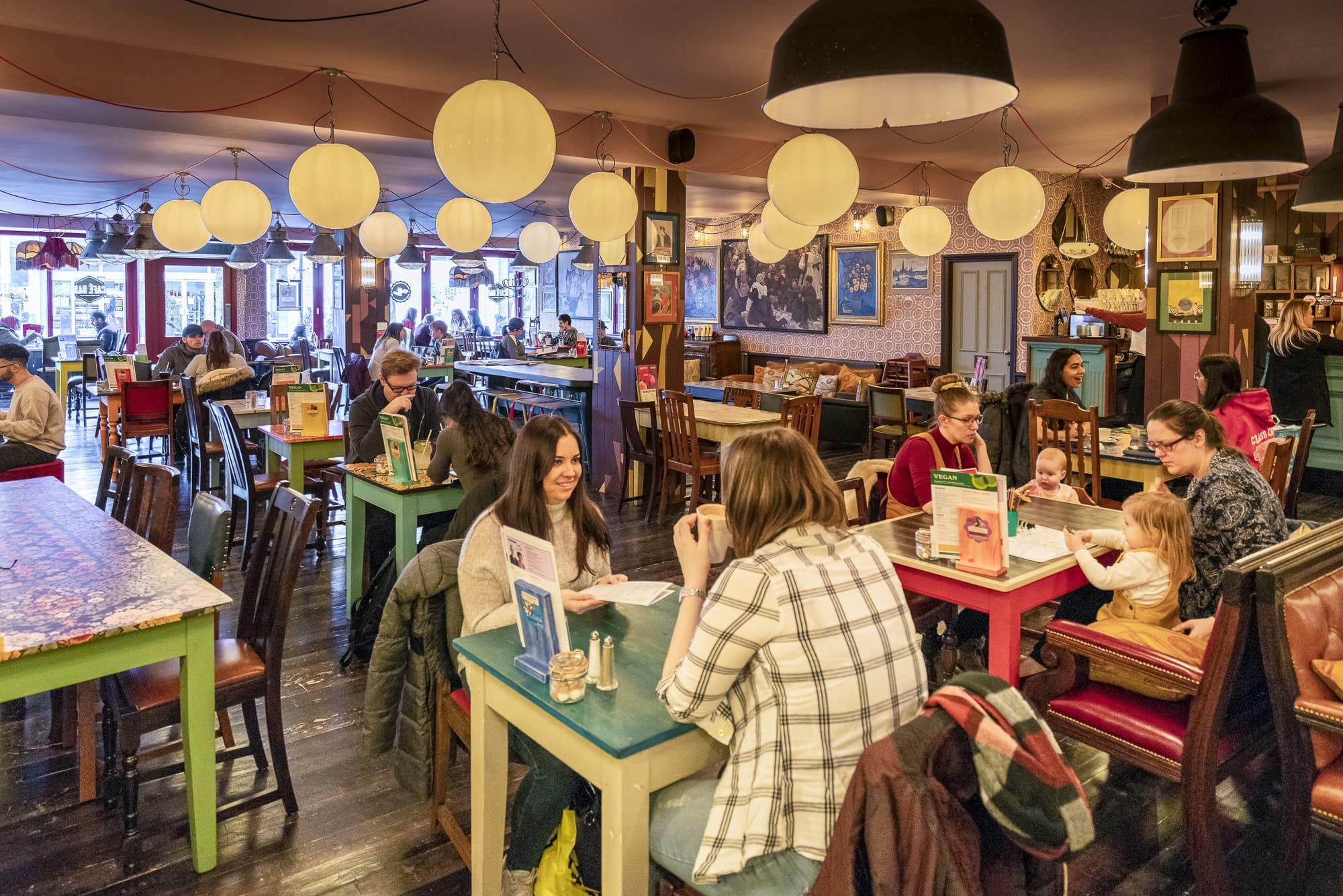 A lively café with colorful tables and chairs, hanging lanterns, and groups of people of various ages chatting, eating, and reading menus in Clio’s warm, bustling atmosphere.