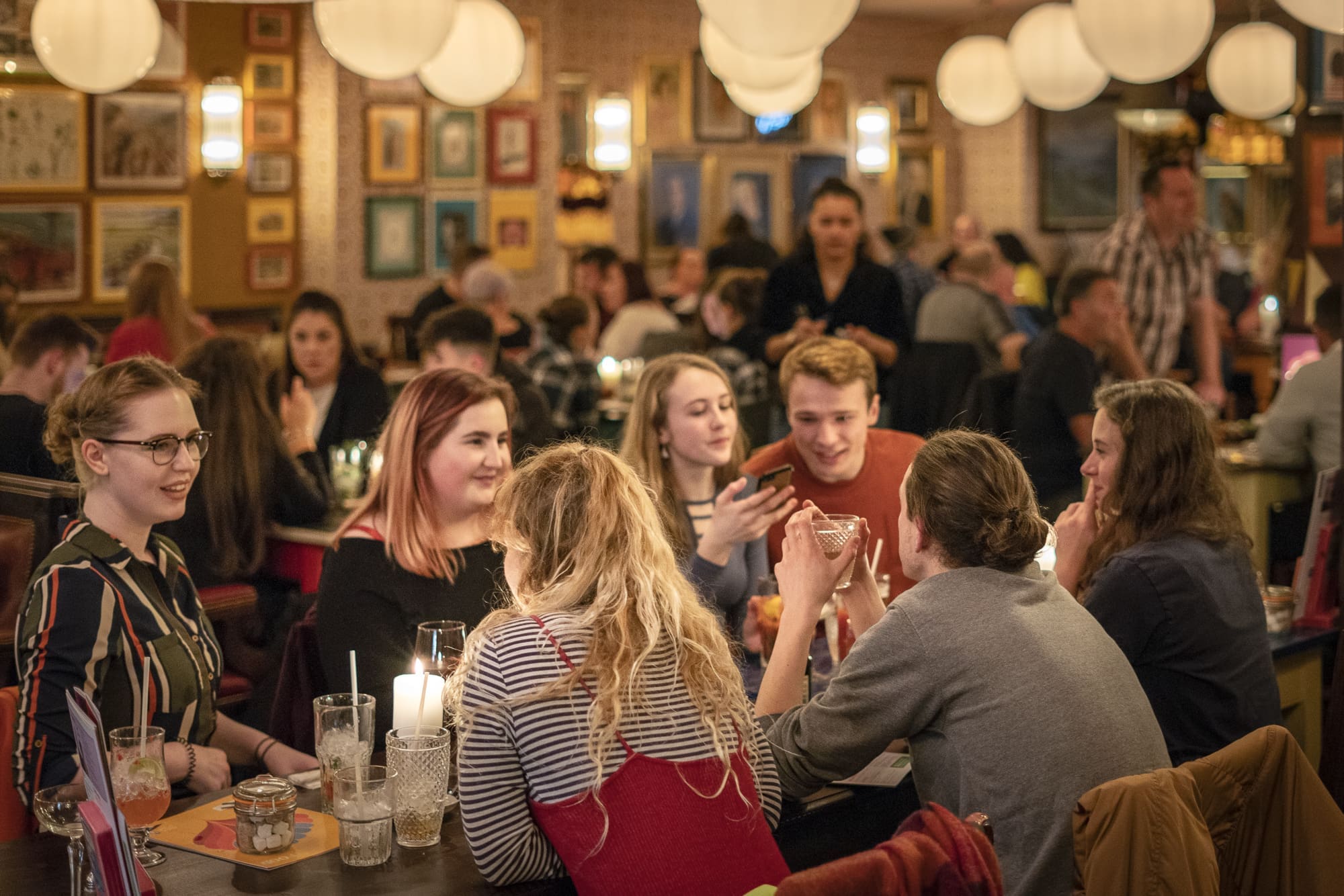 A group of young adults sit around a table in a lively, warmly lit restaurant, talking and enjoying drinks, as Clio captures laughs and conversation with other diners and servers in the background.