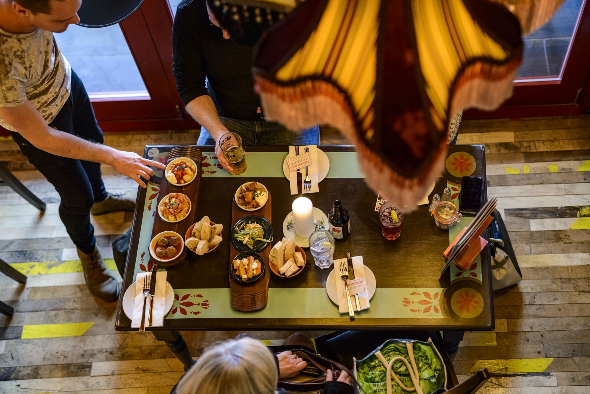 Overhead view of three people at a Panero restaurant table with bowls of food, drinks, and utensils under a large vintage-style lamp. The table sits on a wooden floor with yellow markings.