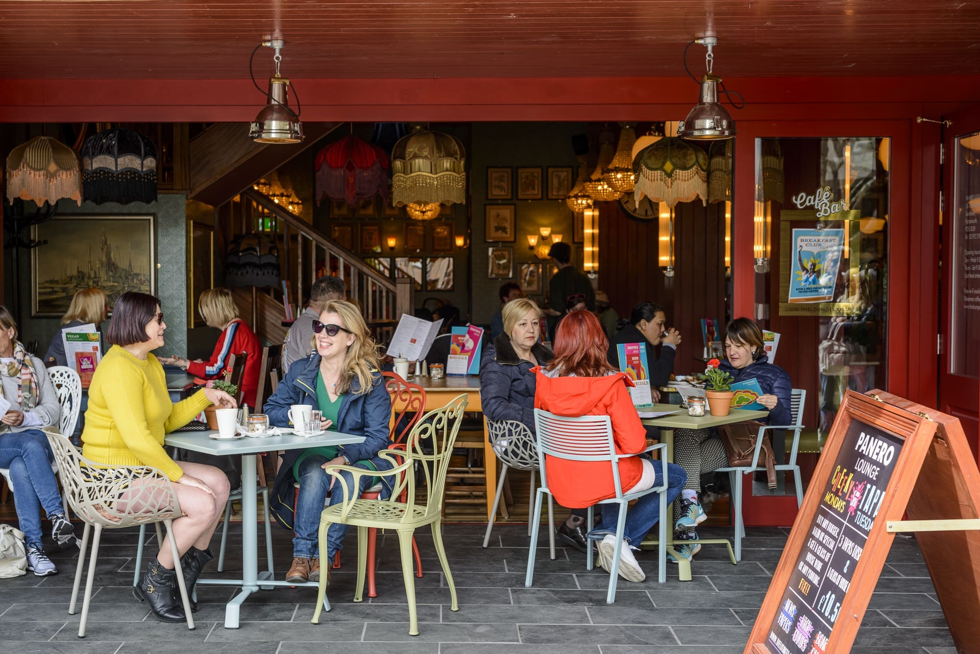 People sit at outdoor tables of Panero, a colorful café, chatting and sipping coffee. Some read menus while others converse. The scene is lively, featuring eclectic decor and a warm, inviting atmosphere in the background.
