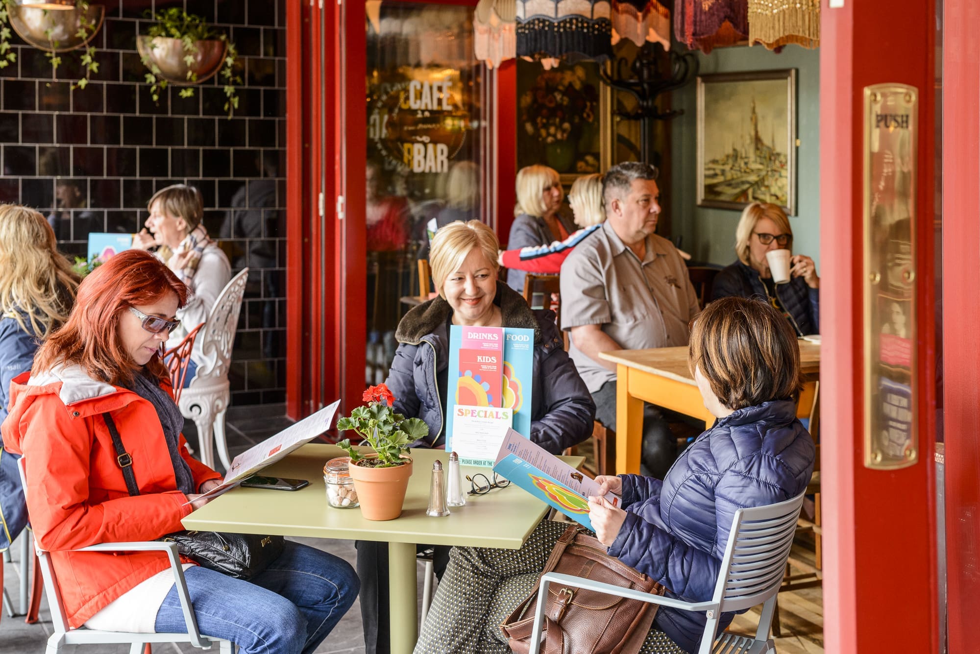 A group of people sit at tables inside the cozy Panero café, reading menus and talking. Potted plants decorate the tables, and a specials sign is displayed. The atmosphere is warm and lively.