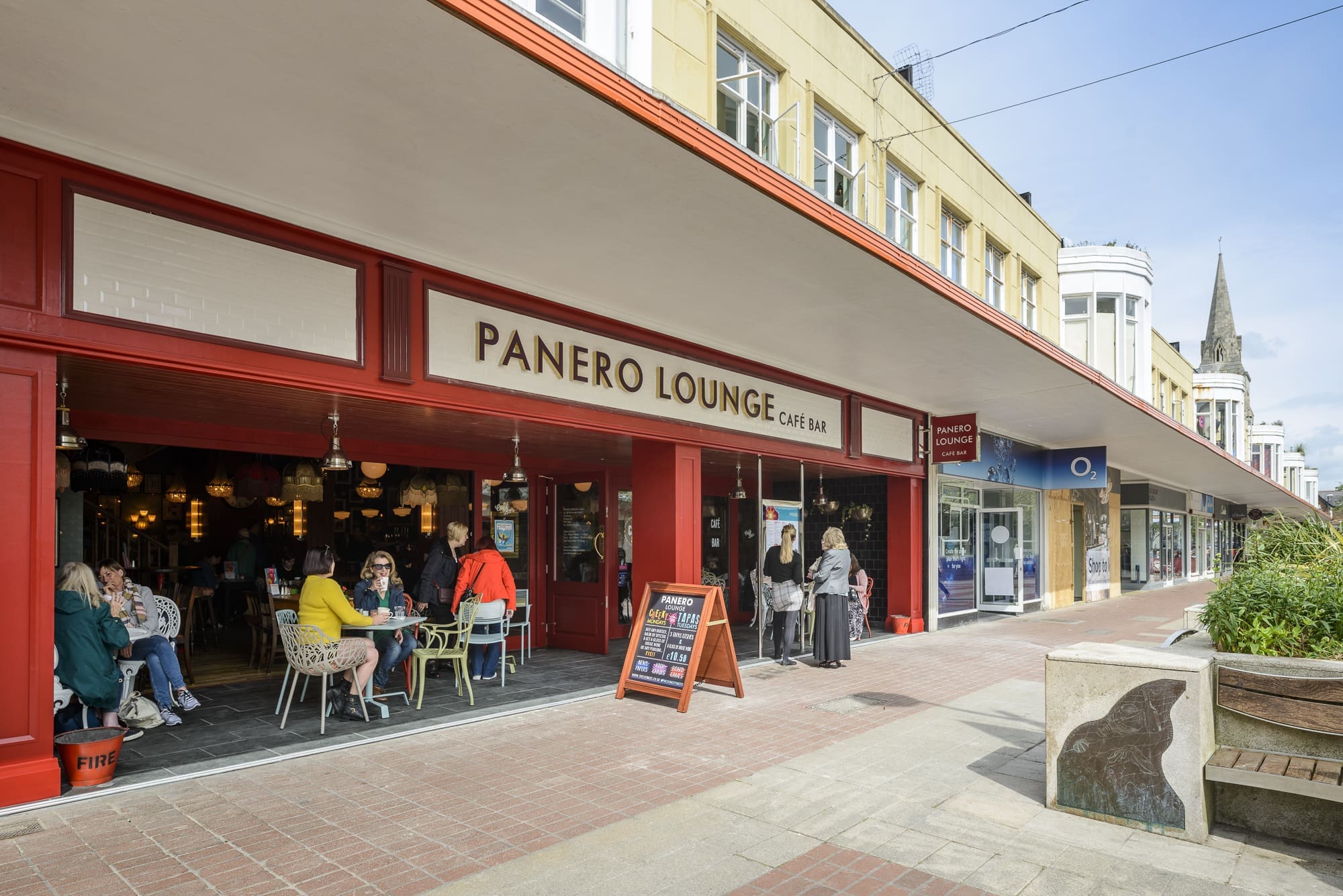 People sit at tables outside Panero Lounge café bar on a pedestrian street with shops. Some customers are eating and talking, while others walk by the lively Panero, marked by its red exterior and large open windows.