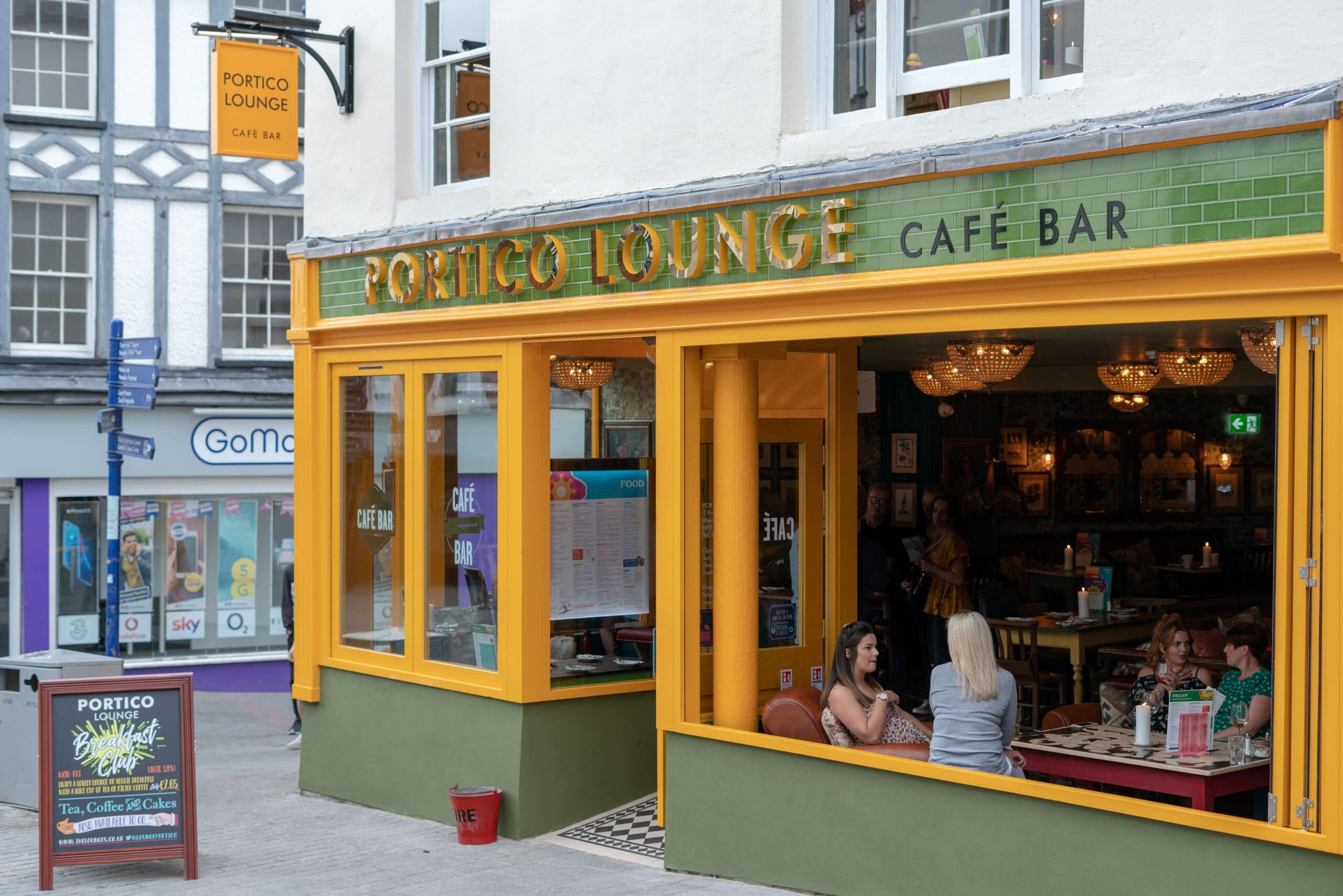 A brightly colored cafe bar named Portico Lounge, with green tiles and yellow trim, features outdoor seating where people are chatting. The warmly lit interior invites guests in, while a sidewalk sign displays the Portico menu.
