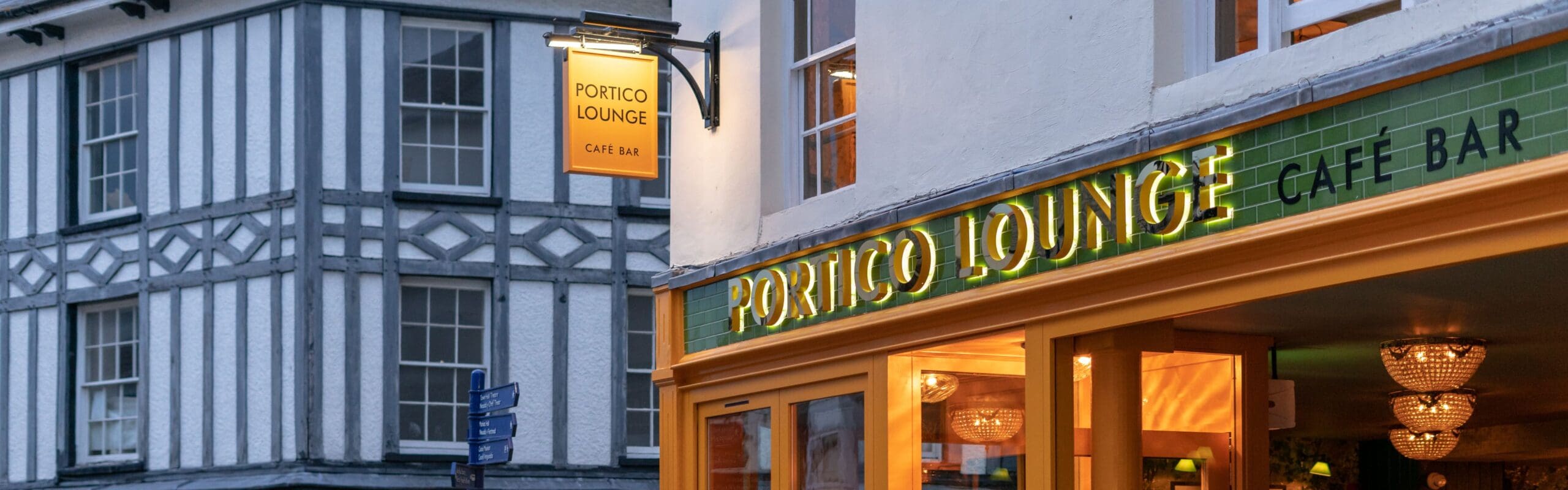 A street view of the Portico Lounge Café Bar with a brightly lit green and yellow Portico sign, beside a timber-framed building with white and gray patterning, in early evening light.