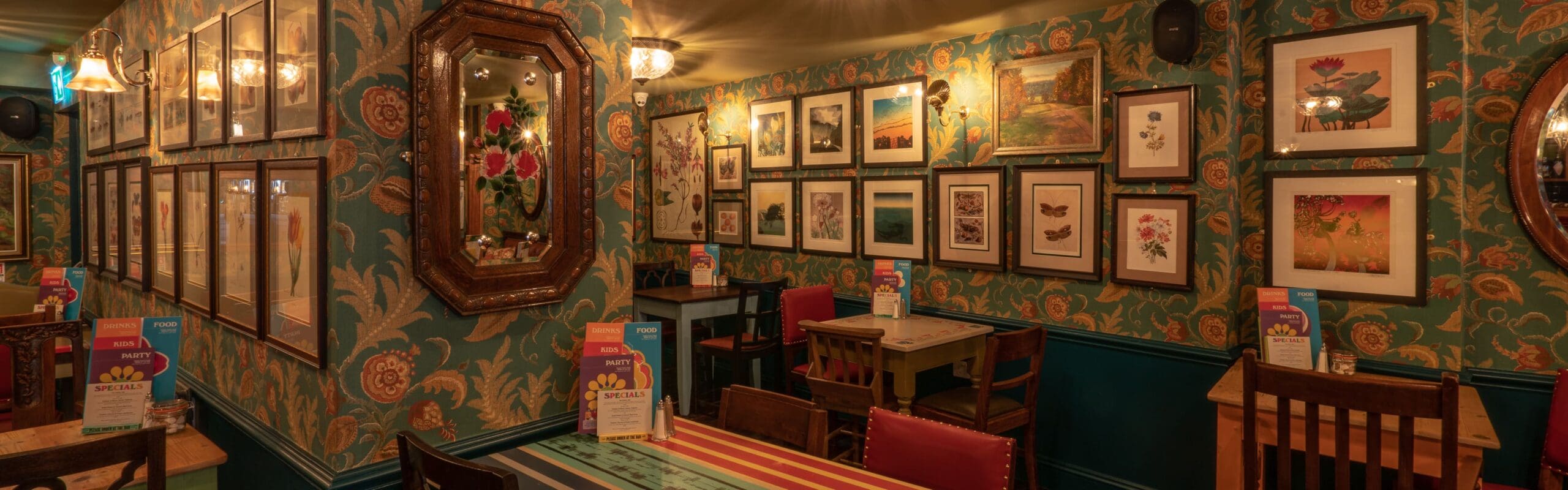 A cozy restaurant interior with a welcoming portico, patterned wallpaper, framed art and mirrors on the walls, wooden tables and chairs, and menu cards on each table, all warmly lit by vintage-style wall lamps.