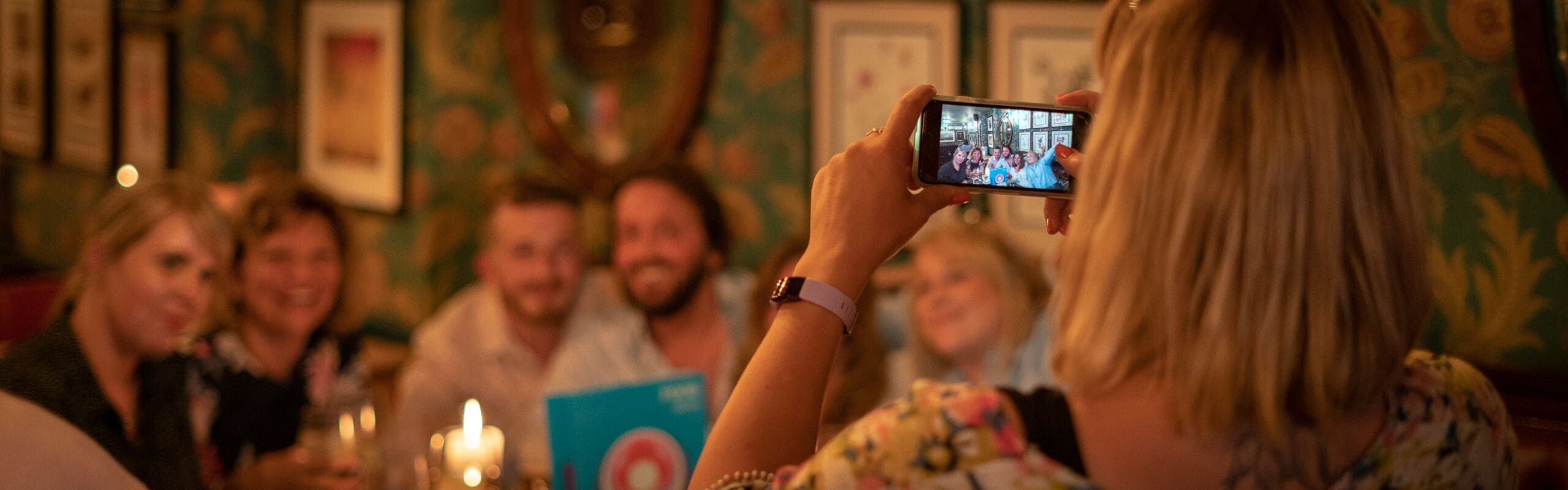 A woman takes a photo with her phone of a group of people smiling and posing together at a dimly lit restaurant with patterned wallpaper, framed pictures, and a view through the portico in the background.
