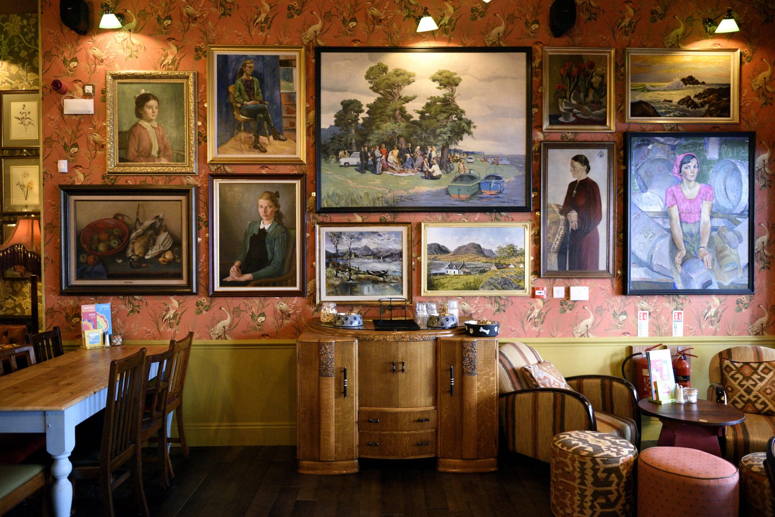 A cozy room with vintage furniture and colorful wallpaper, featuring an Olmo gallery wall of various framed paintings and portraits above a wooden sideboard, dining table, and chairs.