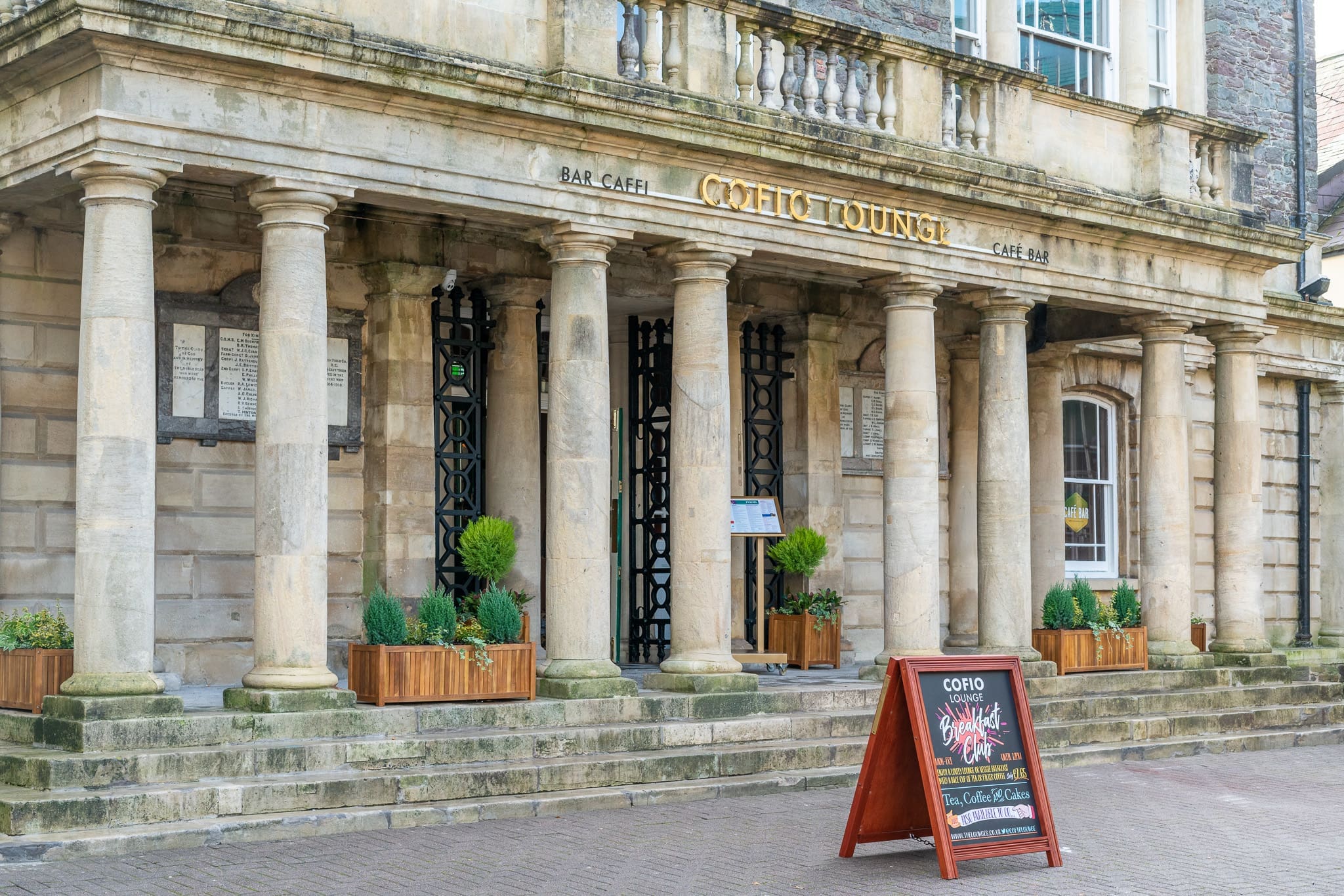 The entrance of a historic stone building with tall columns and a sign reading "Celtic Lounge Cofio." Planters with greenery line the front, and a sandwich board advertises food and drinks on the paved walkway.