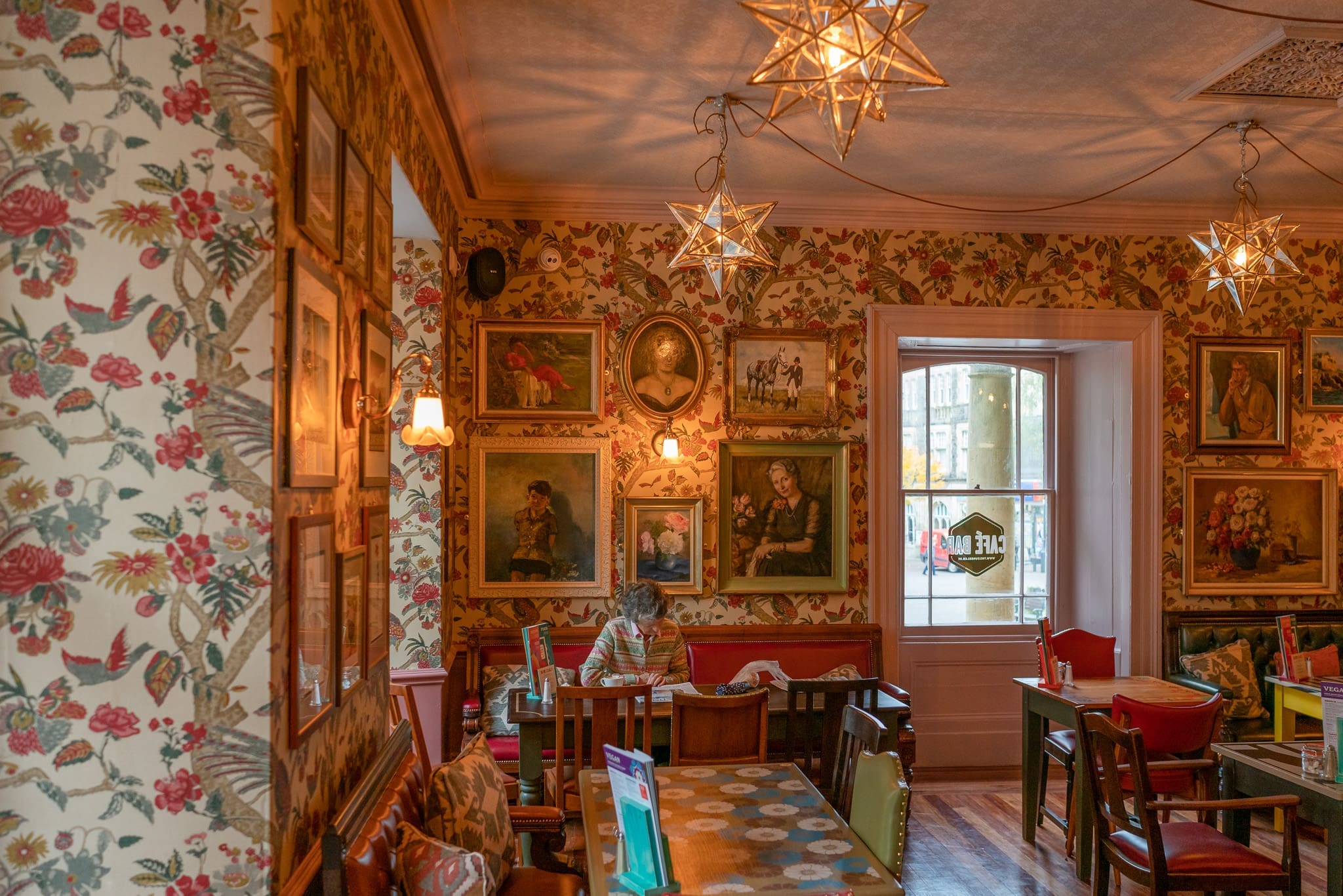 A cozy café with floral wallpaper and vintage framed paintings on the walls. A person sits alone at a table with books, beneath warm Cofio star-shaped lights. Daylight streams in through a window with a view outside.