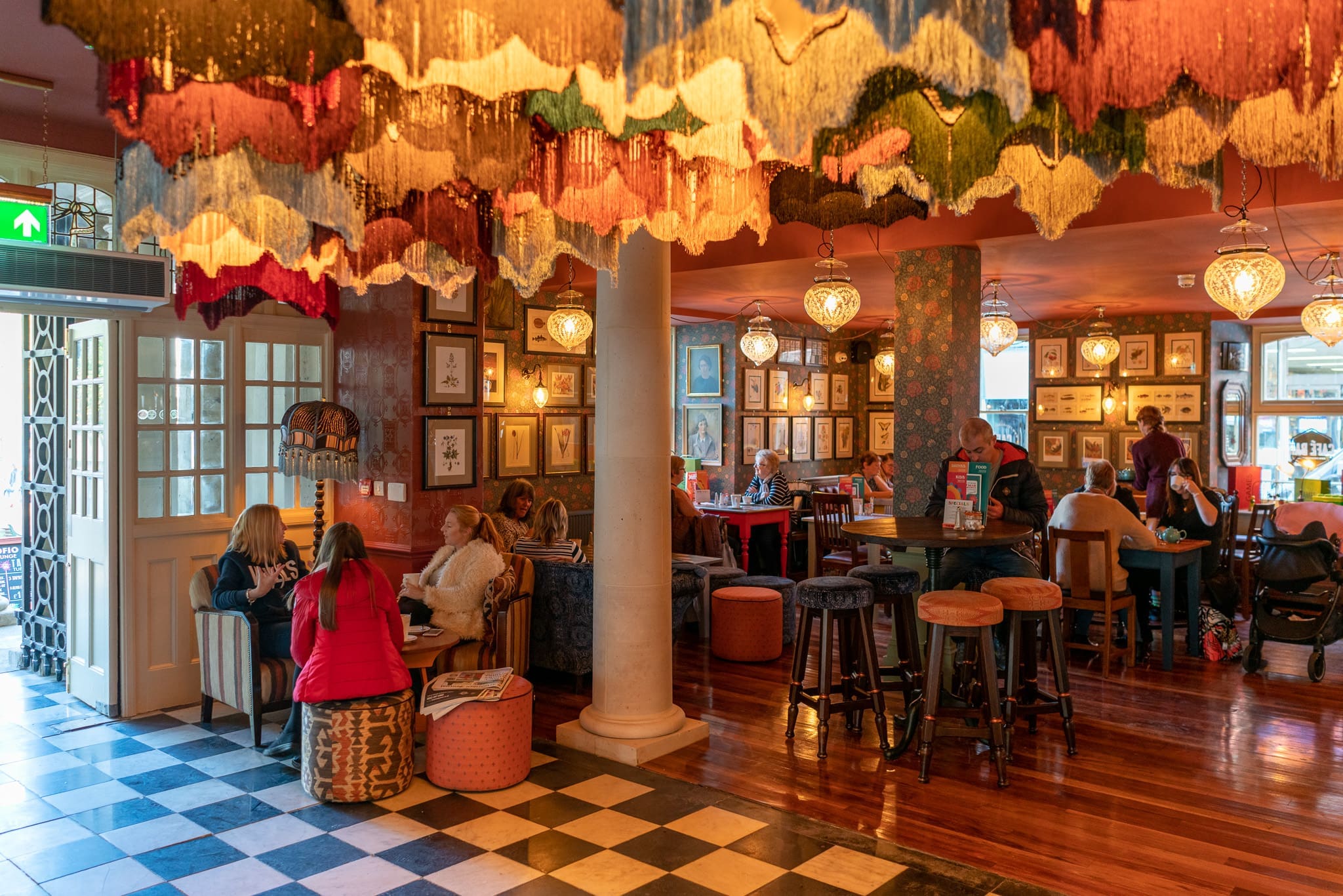 A cozy restaurant interior by Cofio features colorful fringed lamps, framed pictures on the walls, wooden floors, and groups of people chatting at tables and couches while natural light filters in from large windows.