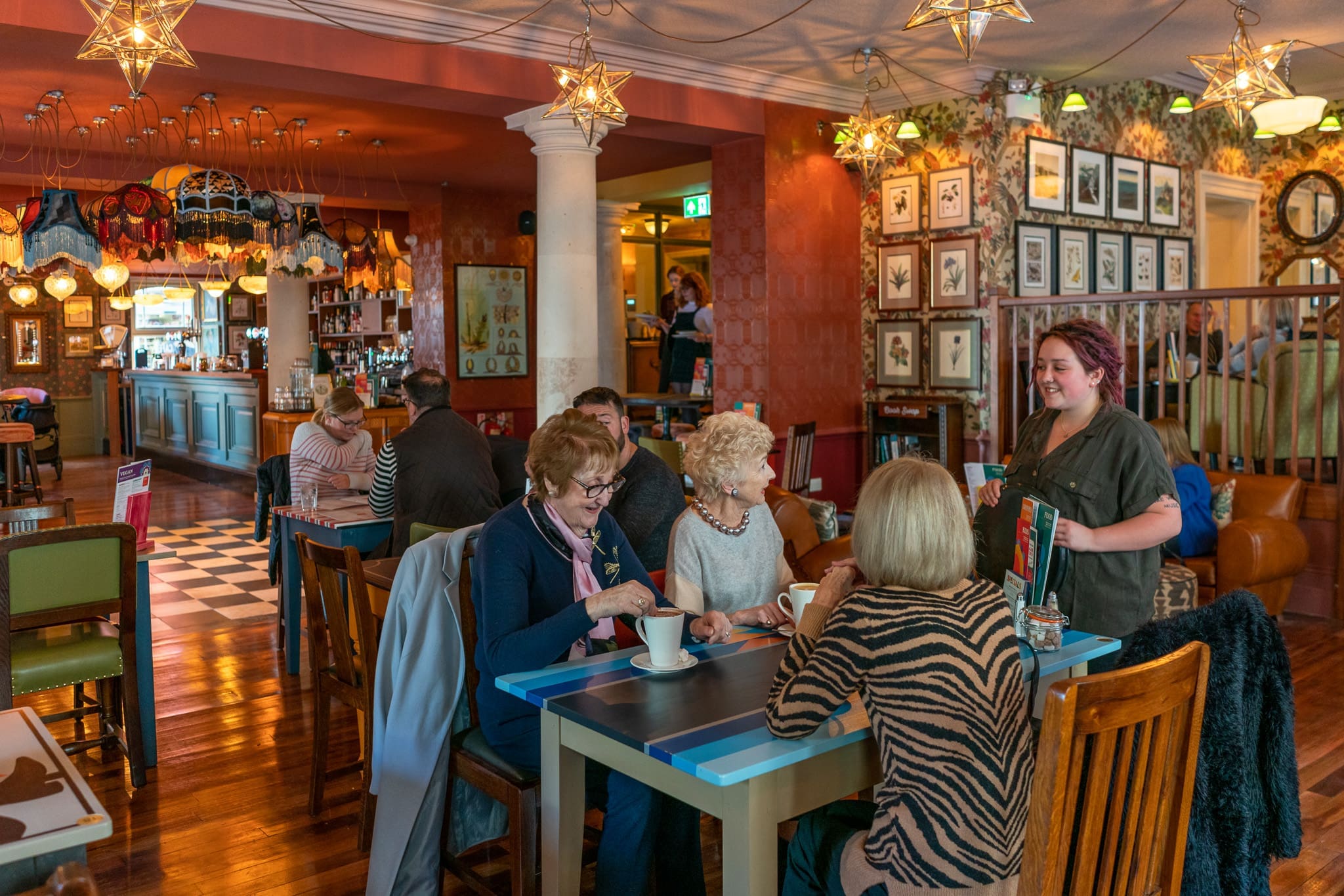 A group of four older women sit at a Cofio café table chatting over drinks; a server stands beside them, smiling. The cozy spot features warm lighting, eclectic decor, and other patrons enjoying the welcoming atmosphere in the background.