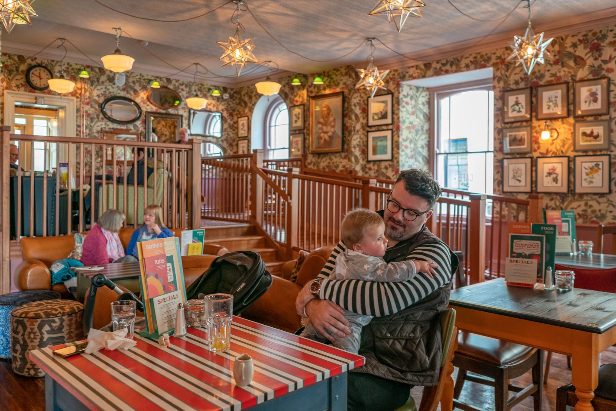 A man in glasses hugs a young child at Cofio, a colorful cafe with patterned wallpaper, star-shaped lights, and framed art. Two women chat on a couch in the background. The atmosphere is cozy and vibrant.