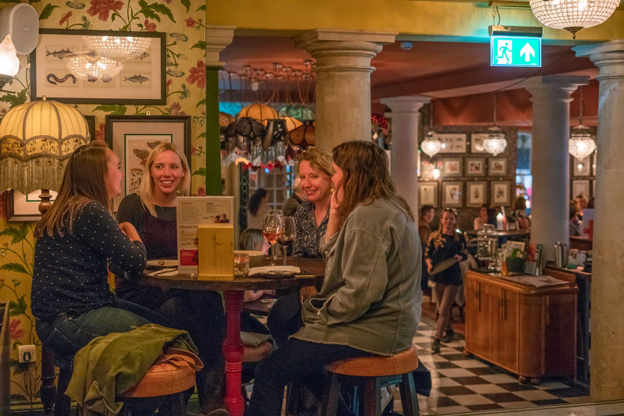 Four women sit around a tall table chatting and drinking at Cofio, a warmly lit, eclectic bar with vintage decor, patterned wallpaper, lamp shades, and framed pictures. Other patrons are visible in the background.