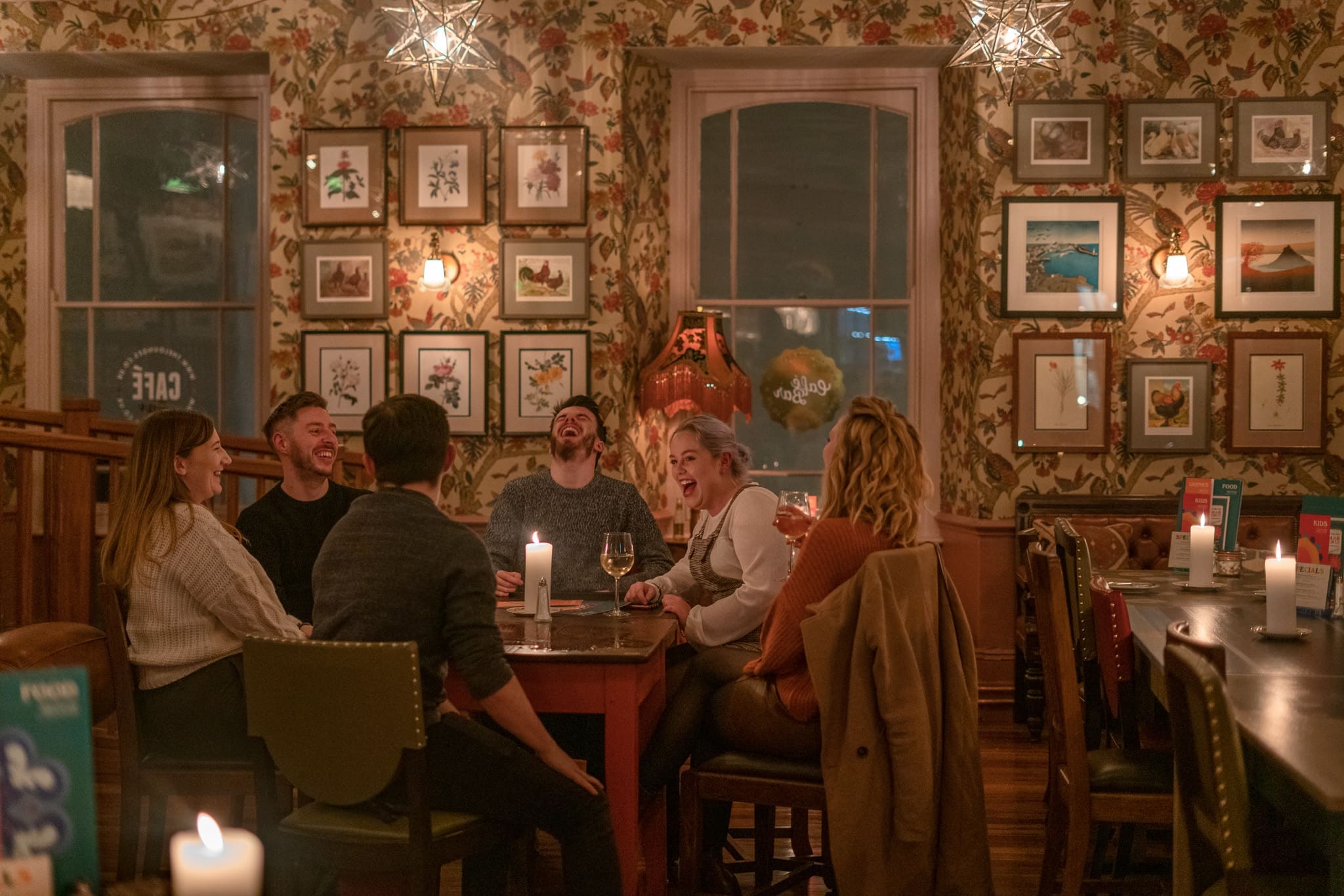 Six people sit around a table in a warmly lit, cozy restaurant like Cofio, laughing together. The floral wallpaper and framed art on the walls, along with candles on the table, create an inviting atmosphere.
