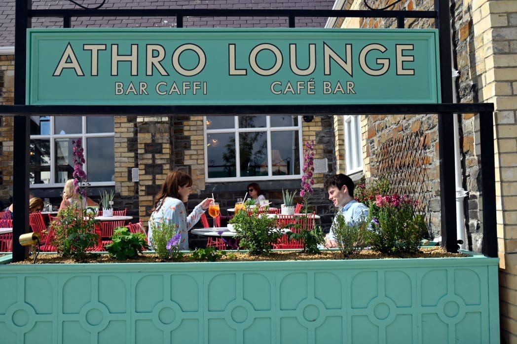 People enjoy drinks at outdoor tables at Athro Lounge café bar. In the foreground, a green sign displays “ATHRO LOUNGE BAR CAFFI CAFÉ BAR.” The lively scene is set against a brick building with plants in the background.
