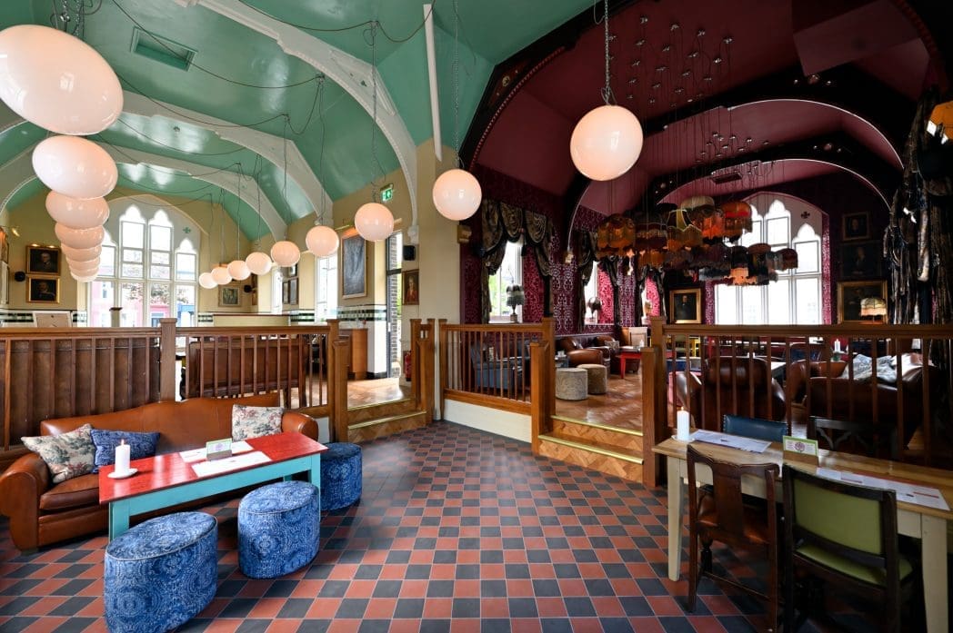 A spacious, colorful Athro lounge with high mint green ceilings, round hanging lights, leather sofas, patterned cushions, wooden railings, and checkered tile flooring leads to a cozy, warmly lit seating area with arched decor.